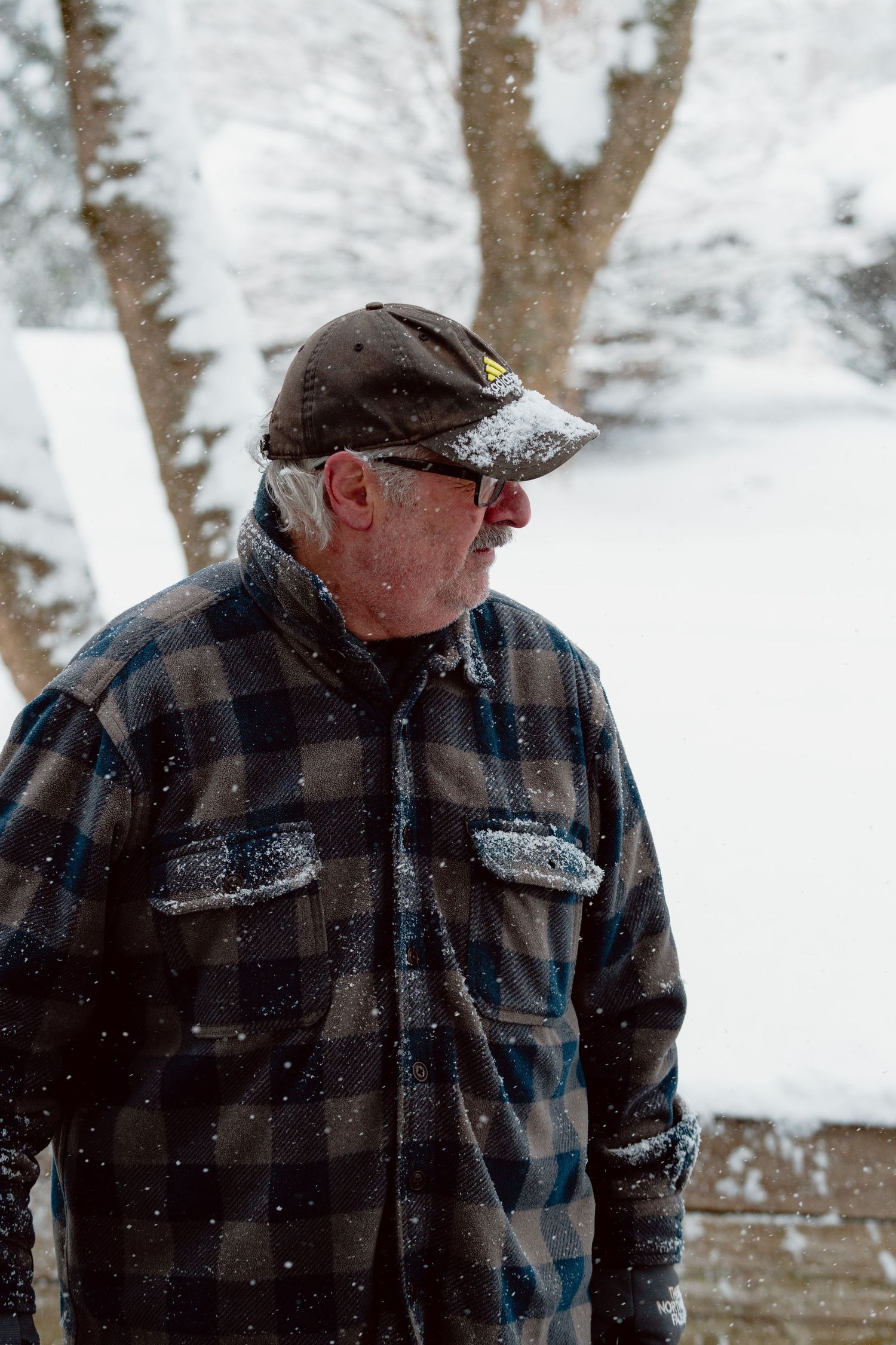 Elderly man in plaid jacket and cap looking sideways while standing in falling winter snow.