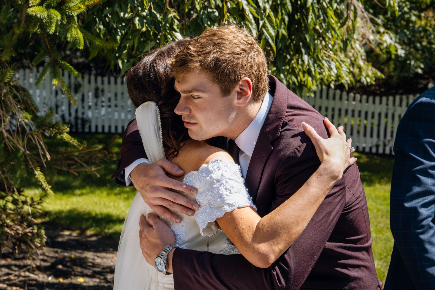 A couple shares a tender embrace in wedding attire outdoors, with lush greenery and a white fence in the background.
