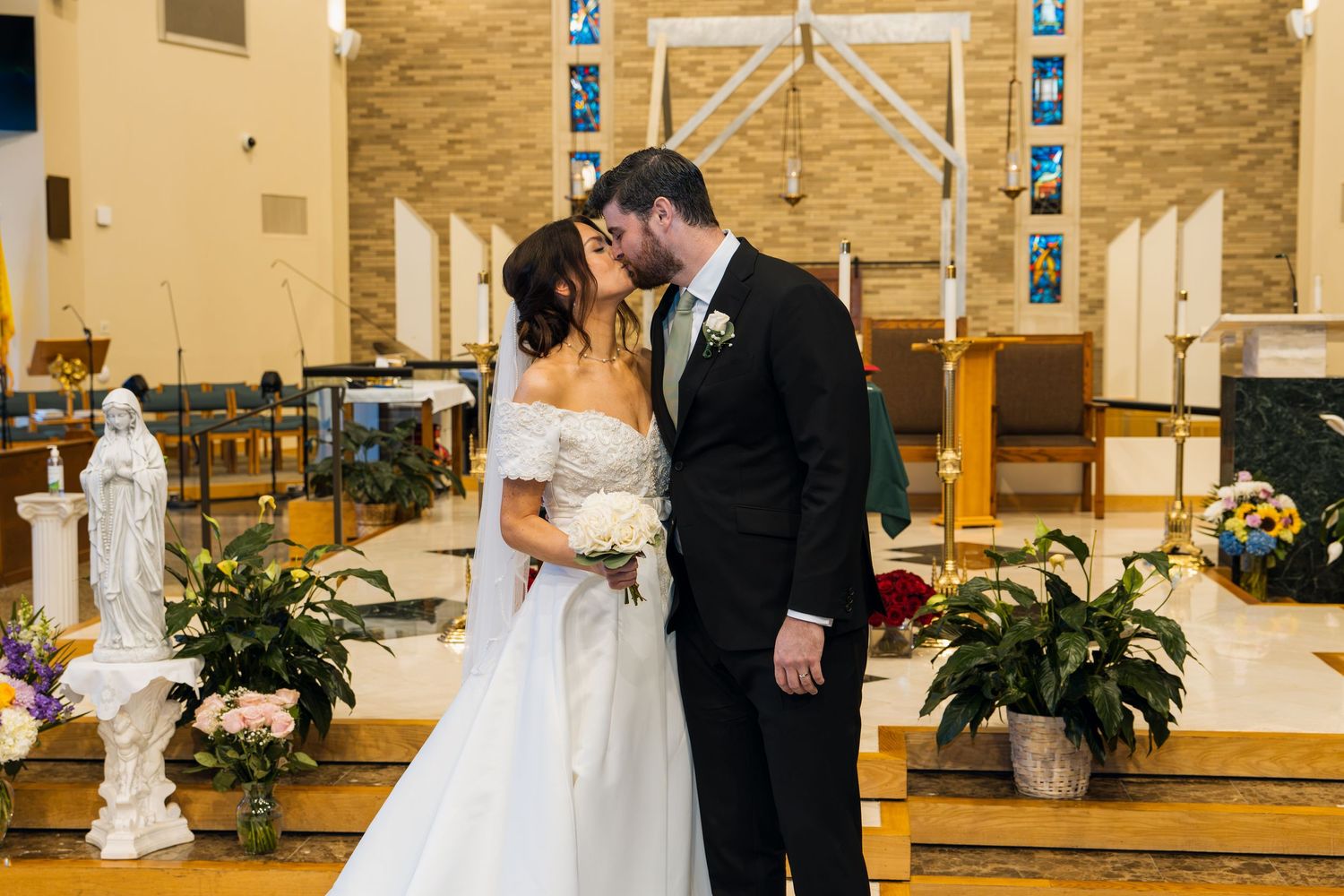 Bride and groom share a kiss at church altar, bride in off-shoulder gown holding bouquet, groom in black suit.