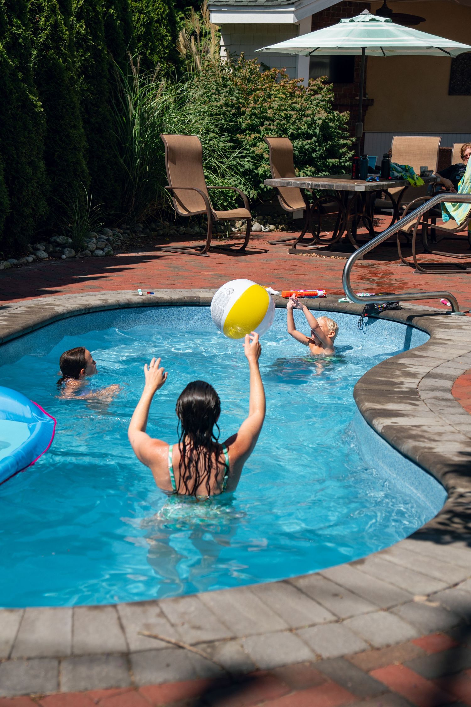 Woman tossing a yellow and white beach ball to children in a sunny backyard pool.