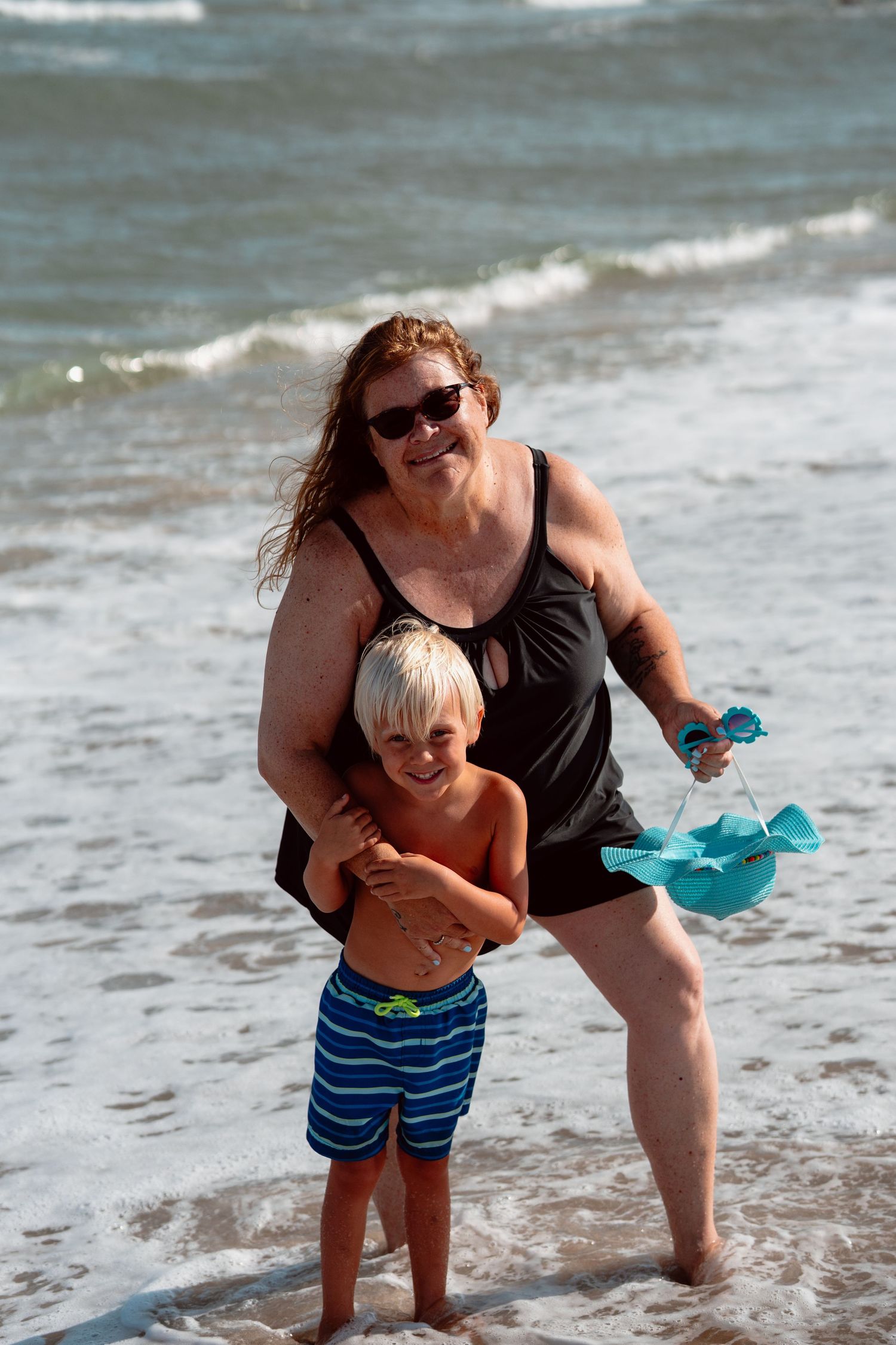 Happy grandmother hugging young blonde boy in blue swimsuit while standing in ocean waves.