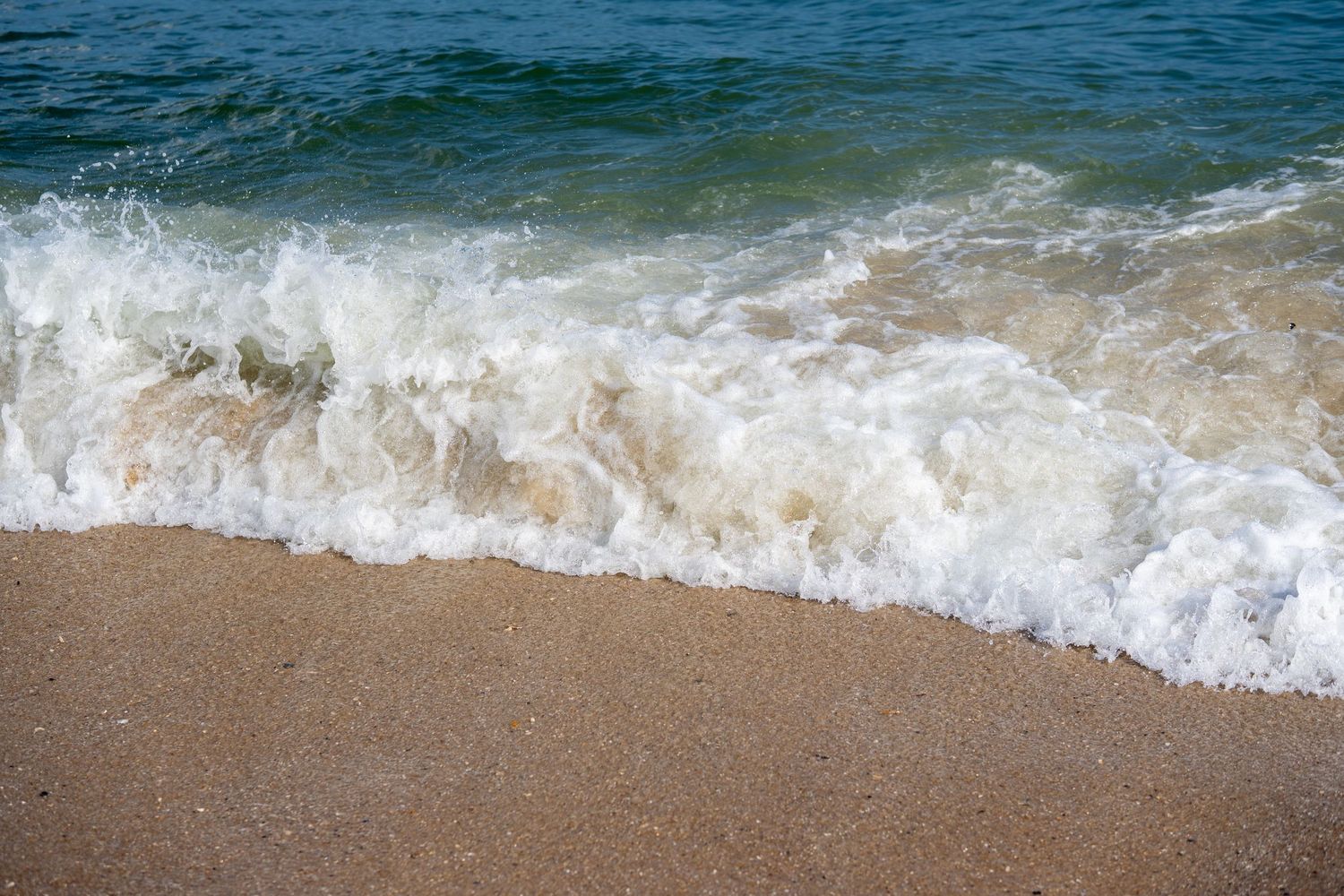White foamy ocean wave rolling onto a sandy beach shore with clear blue-green water.