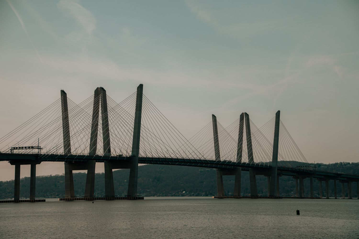Cable-stayed bridge with tall pylons spans a wide calm river under a pale overcast sky.
