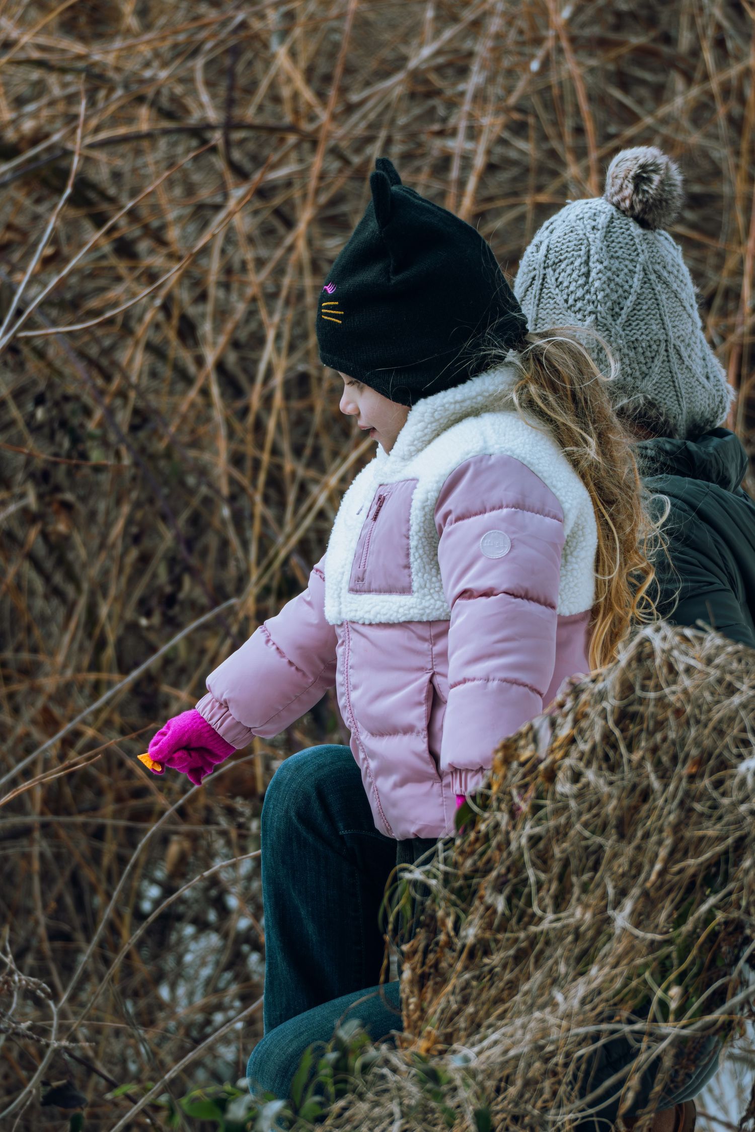 Young child in pink jacket and black cat hat playing outdoors in winter brush.