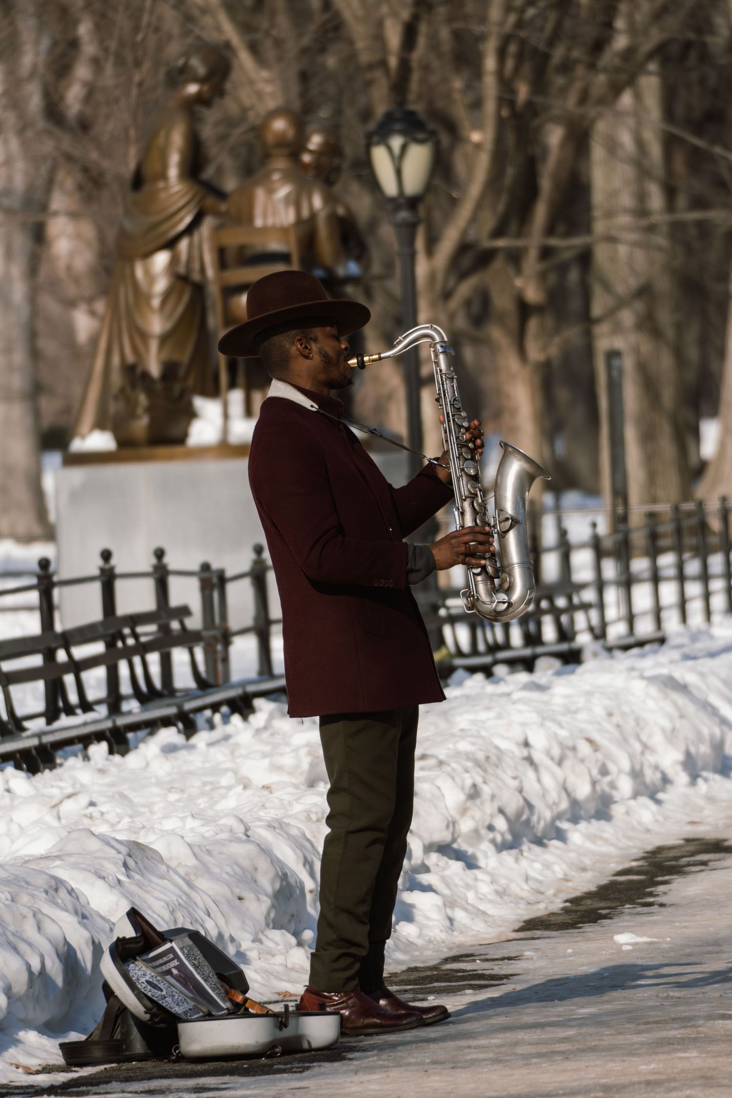 A street musician in a burgundy blazer and brown hat plays saxophone in a snowy Central Park, NYC.