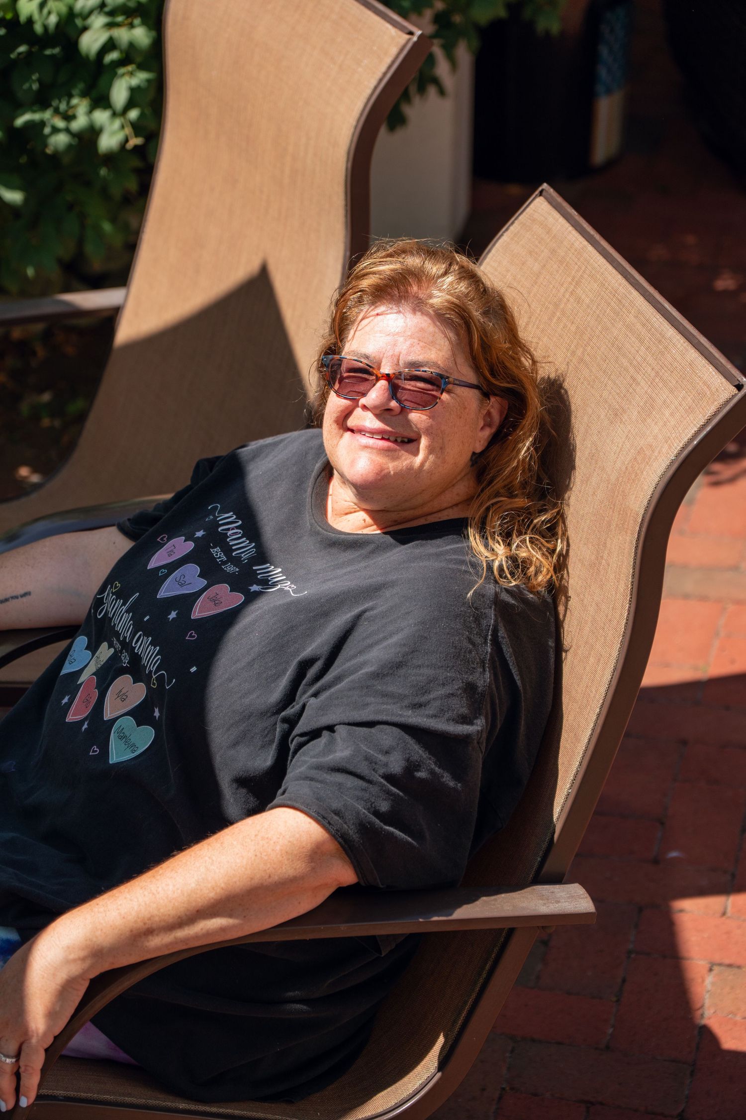 Smiling middle-aged woman with red hair relaxes in outdoor patio chair on sunny day.