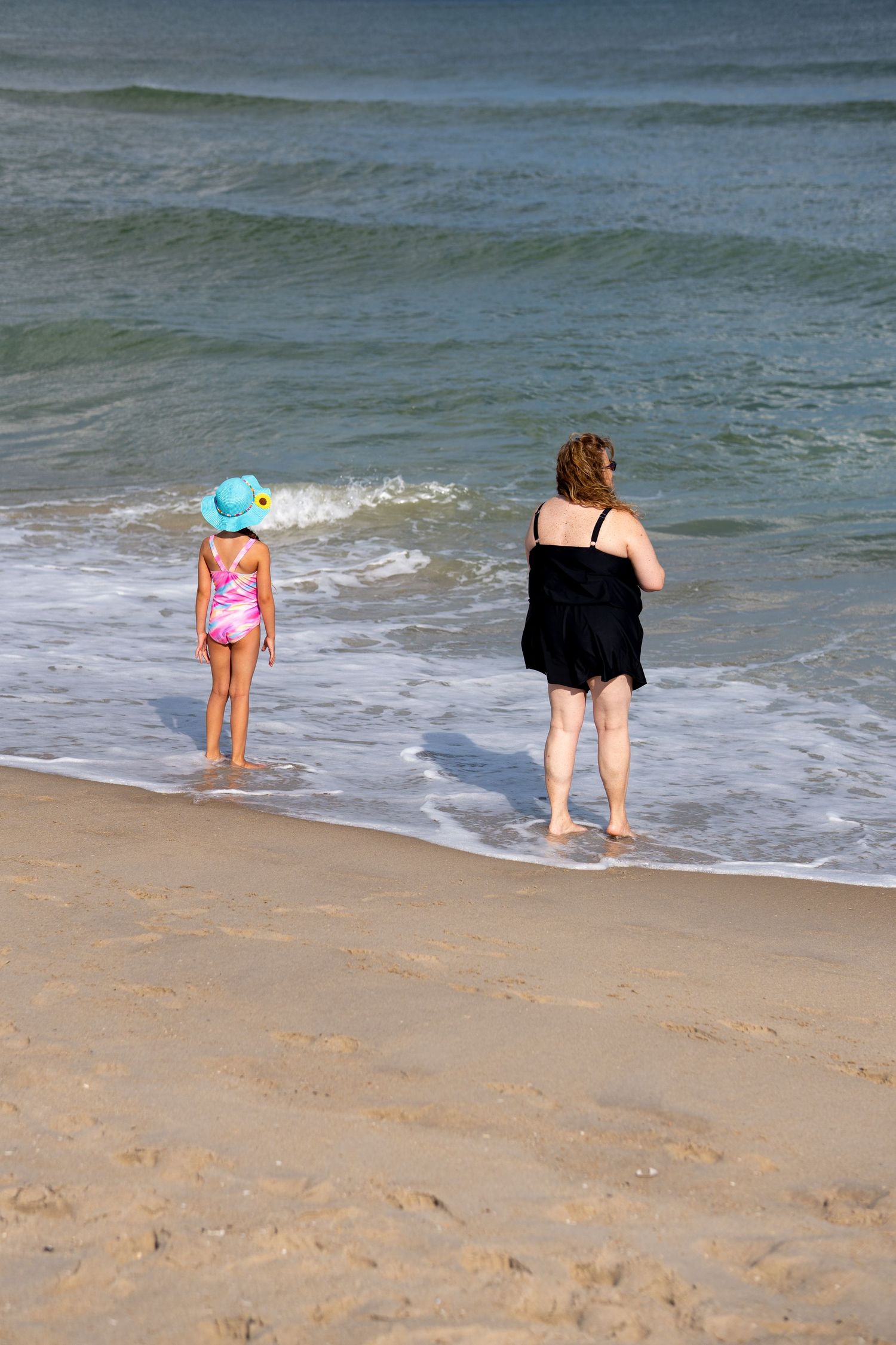 Woman and young girl in blue hat wading at the edge of ocean waves on sandy beach.
