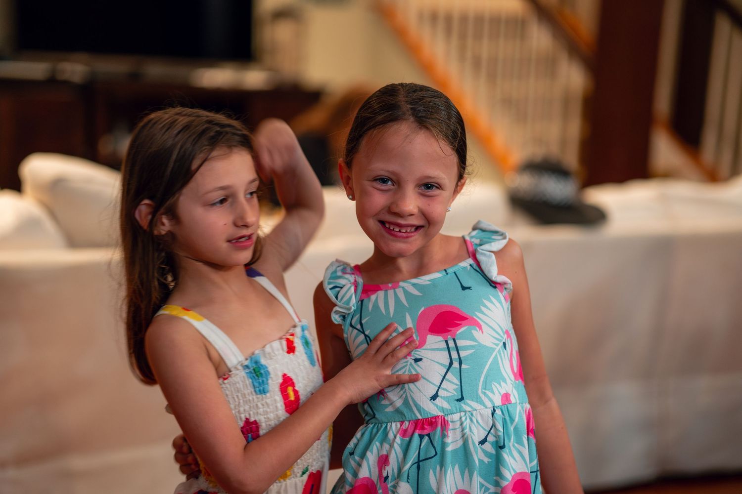Two young girls in summer dresses smiling together in a warmly lit living room.