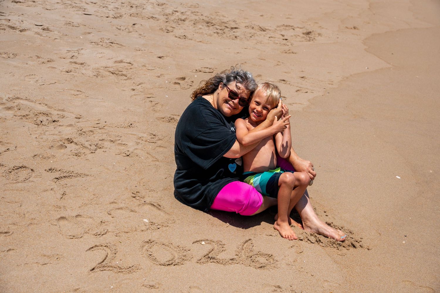 Woman and child hugging on sandy beach with 2026 written in sand on a sunny day.