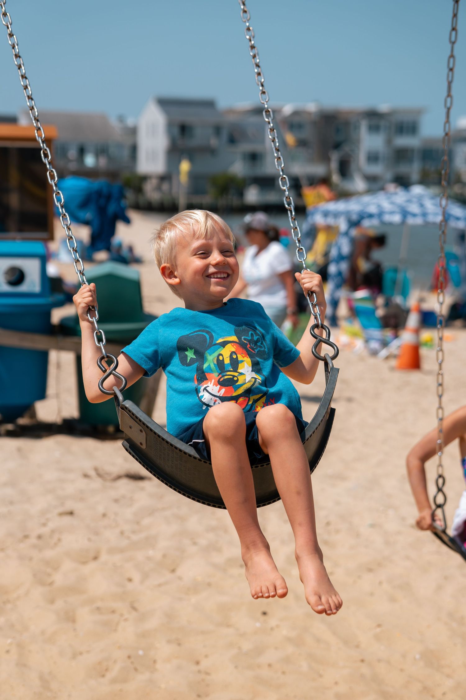 Happy blonde boy in Mickey Mouse shirt swinging on beach playground swing on sunny day.