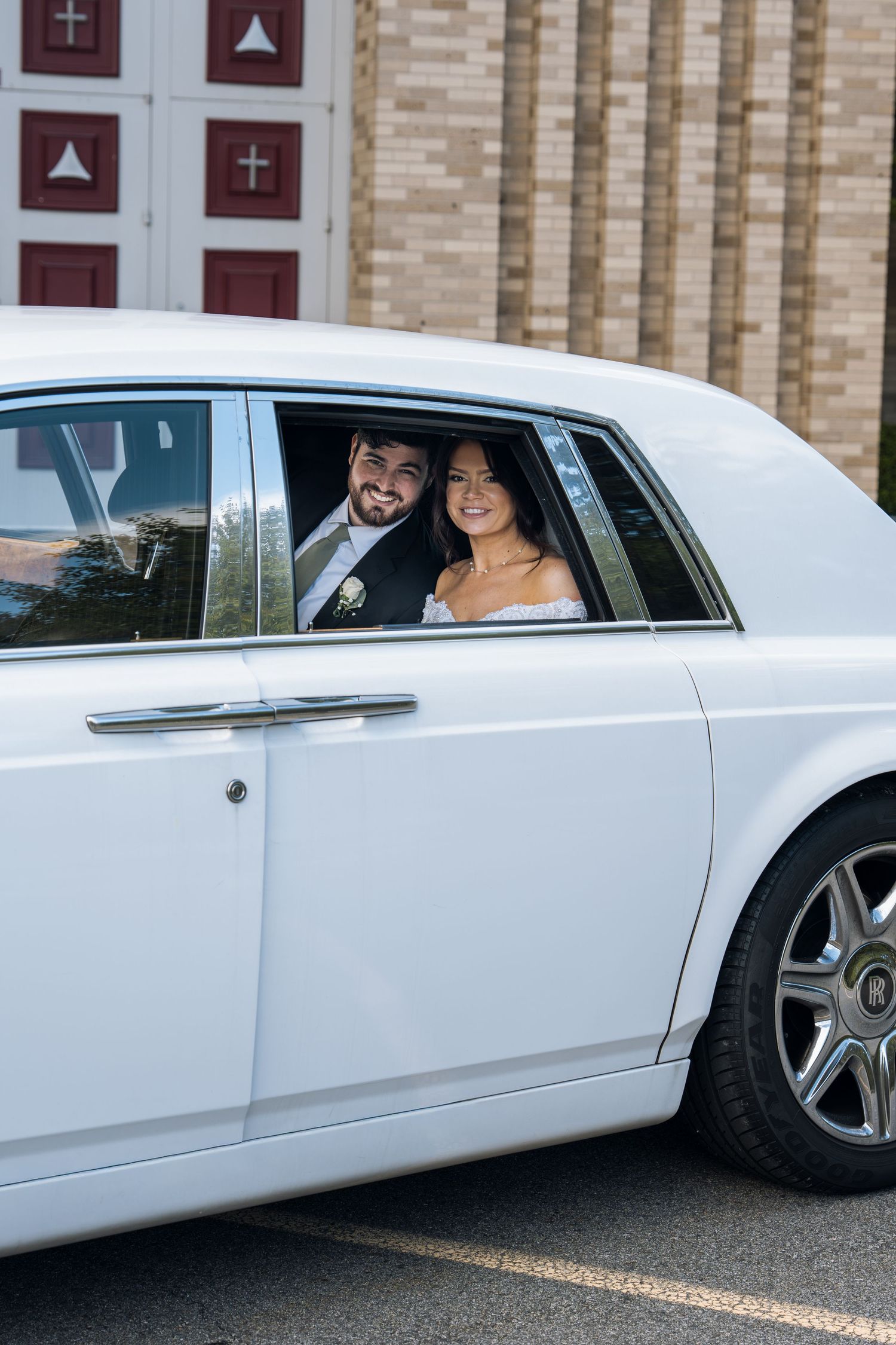 Smiling bride and groom in wedding attire seated inside a white luxury car outside a church.