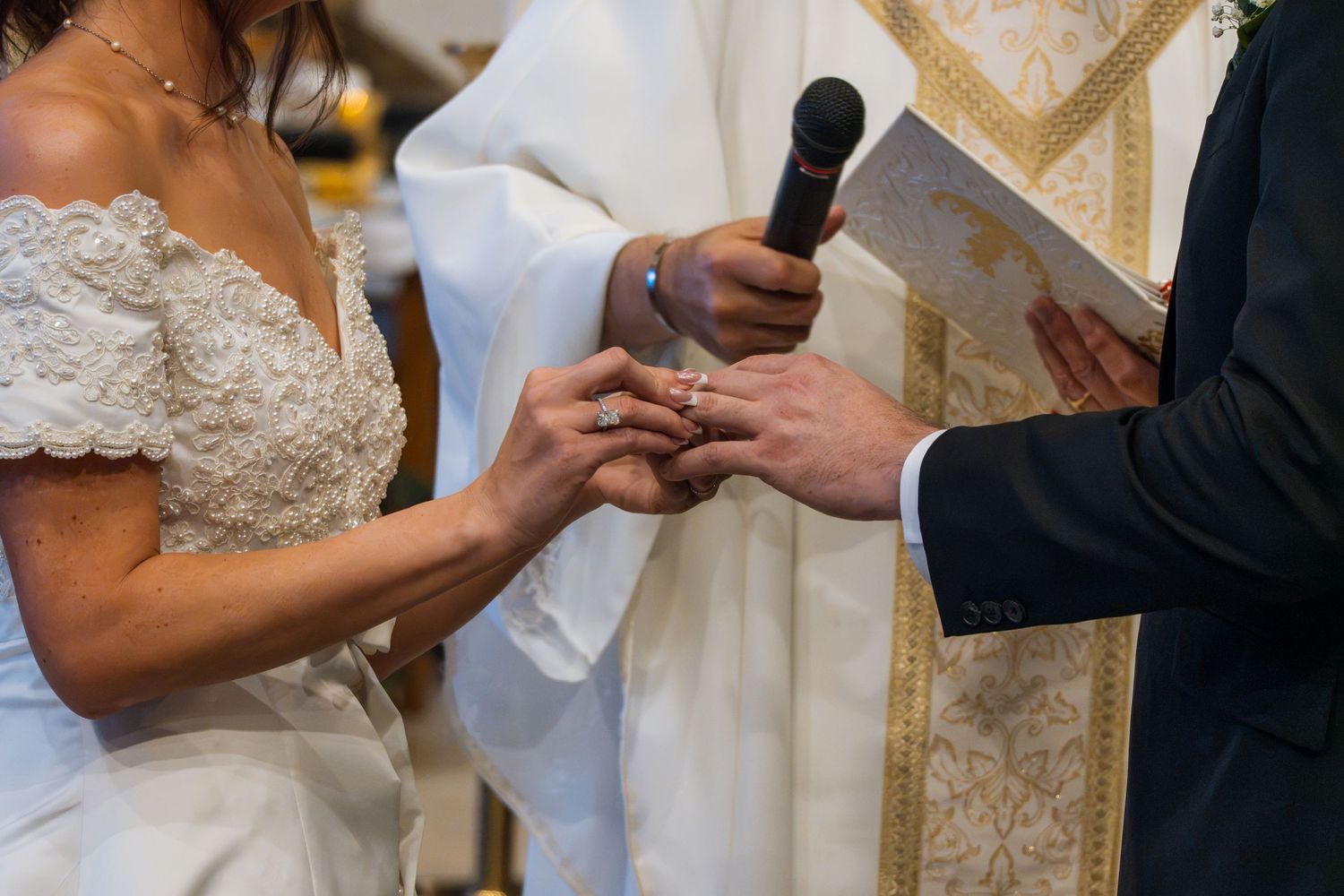 Groom placing a wedding ring on bride's finger during a church ceremony, officiated by a priest holding a microphone.