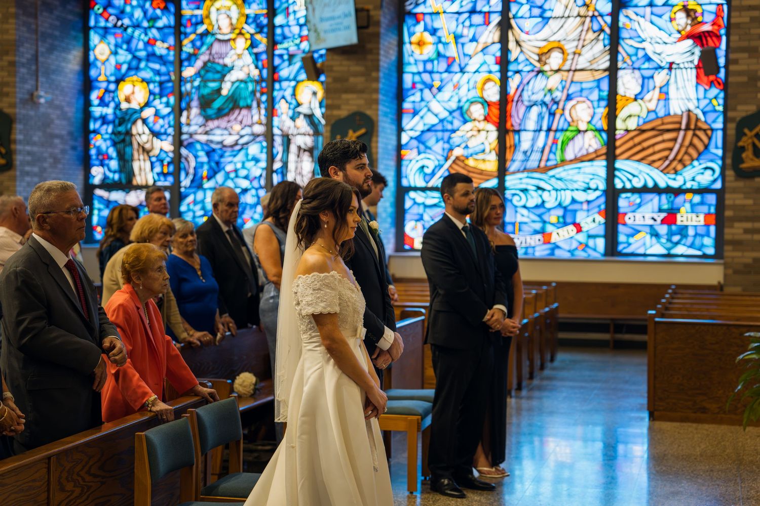 Bride in white gown walks down church aisle with stained glass windows and seated guests during wedding ceremony.