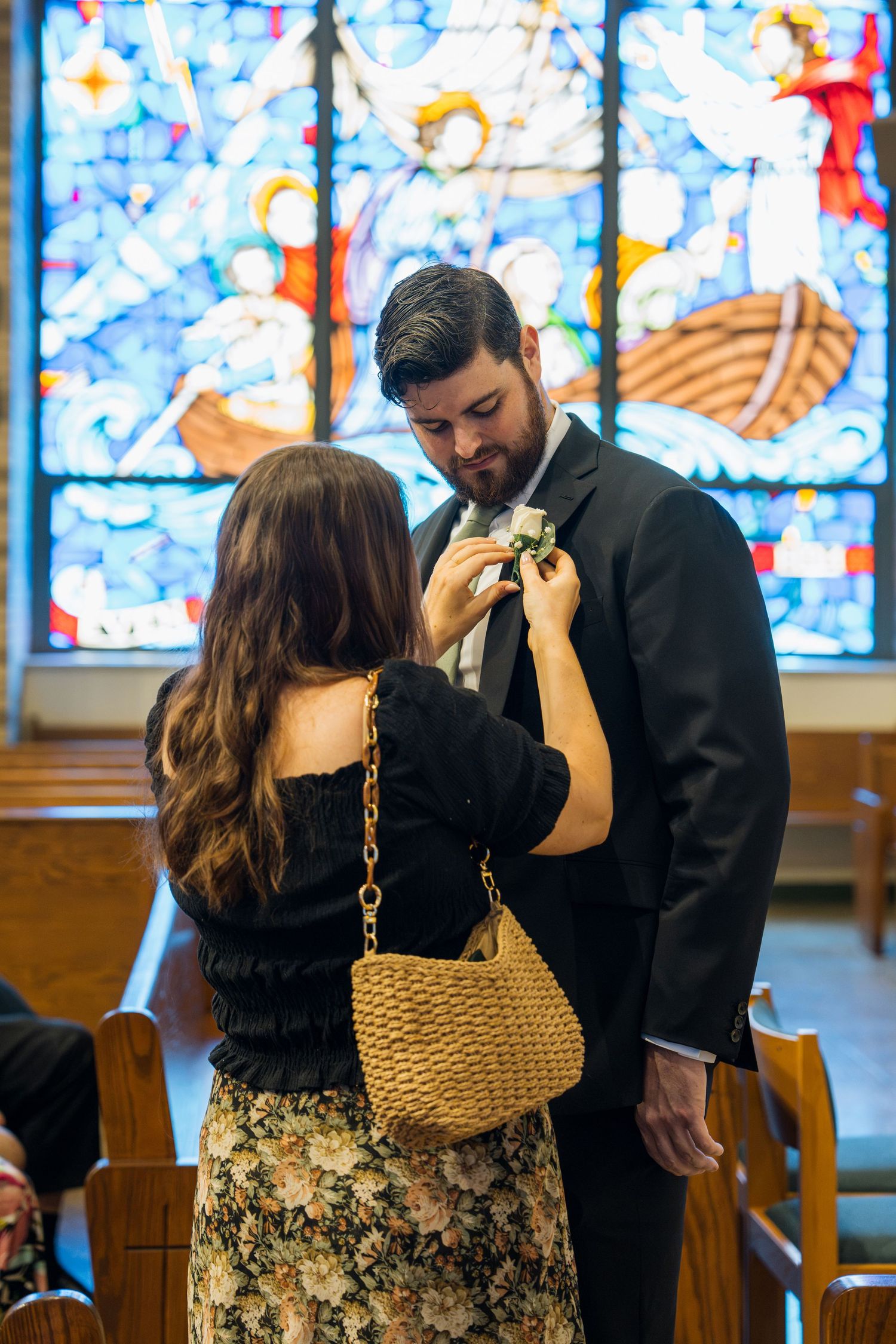 Woman pinning boutonniere on man in black suit before a colorful stained glass window in a church.