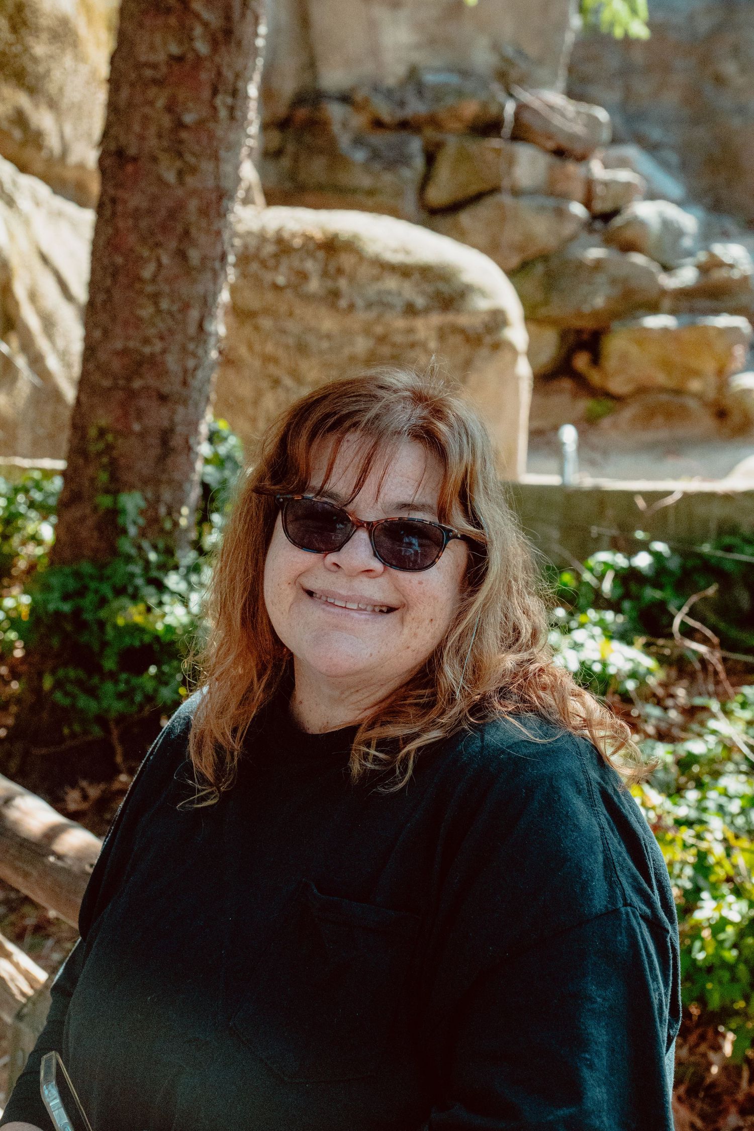 A smiling woman with sunglasses and auburn hair posing outdoors near trees and rocks.