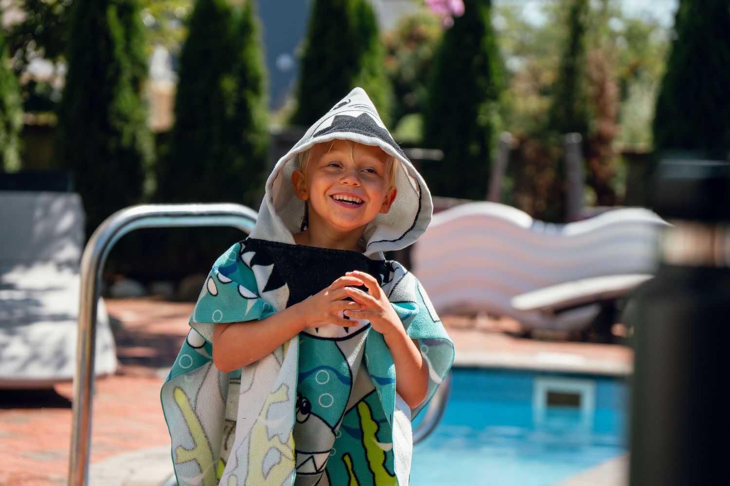 Happy child smiling in a colorful shark hooded beach towel by a sunny backyard pool.