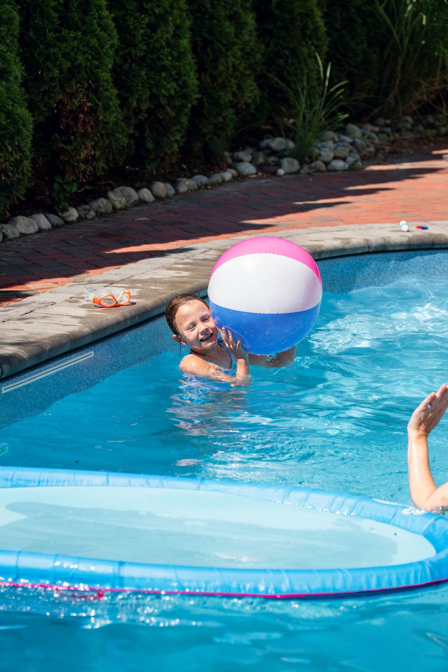 Child playing with a large colorful beach ball in a sunny backyard swimming pool.
