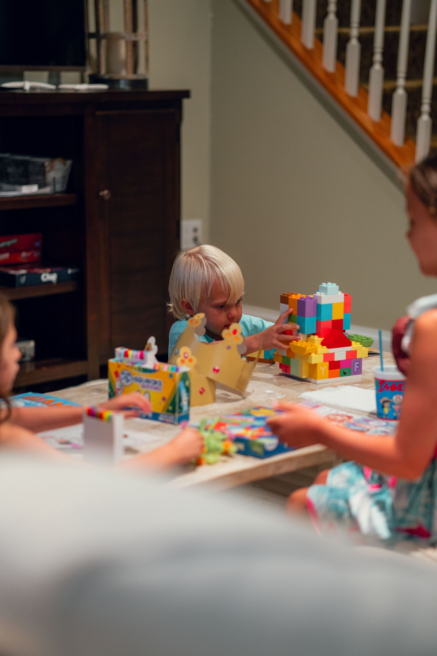 Young blonde child focused on building with colorful blocks at a coffee table with siblings nearby.