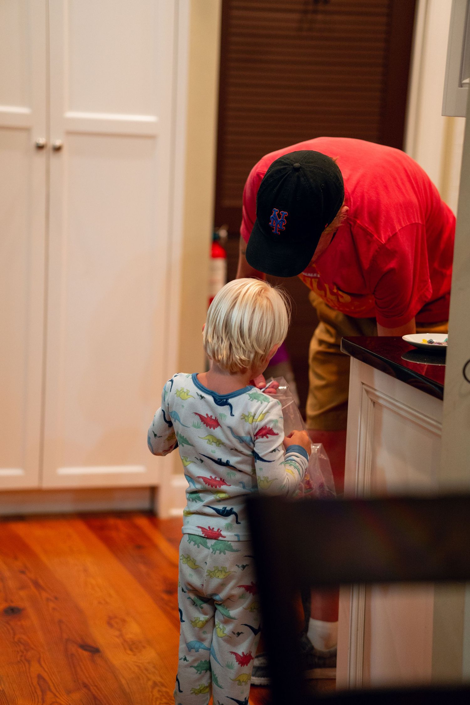 A toddler in dinosaur pajamas standing beside an adult in a kitchen.