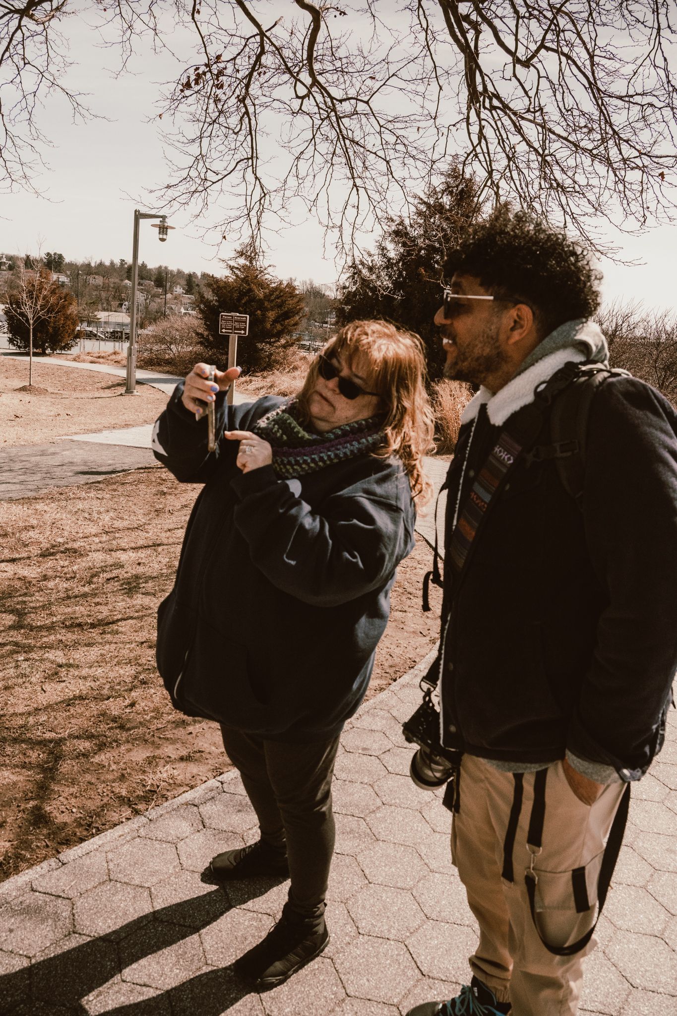 Two people in winter coats examine a wooden sign outdoors on a sunny day near bare trees.