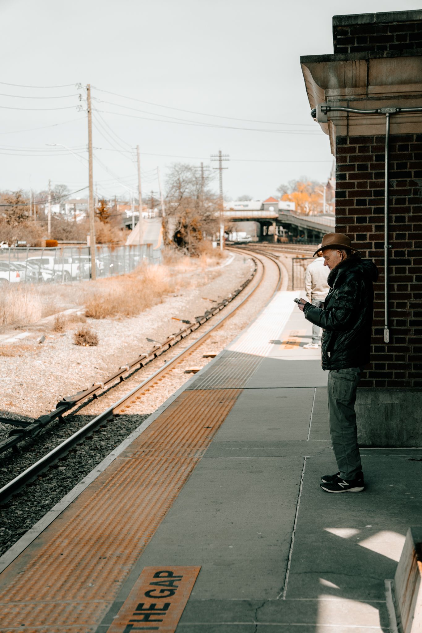 Person in black jacket standing on an empty train platform beside brick building checking their phone.