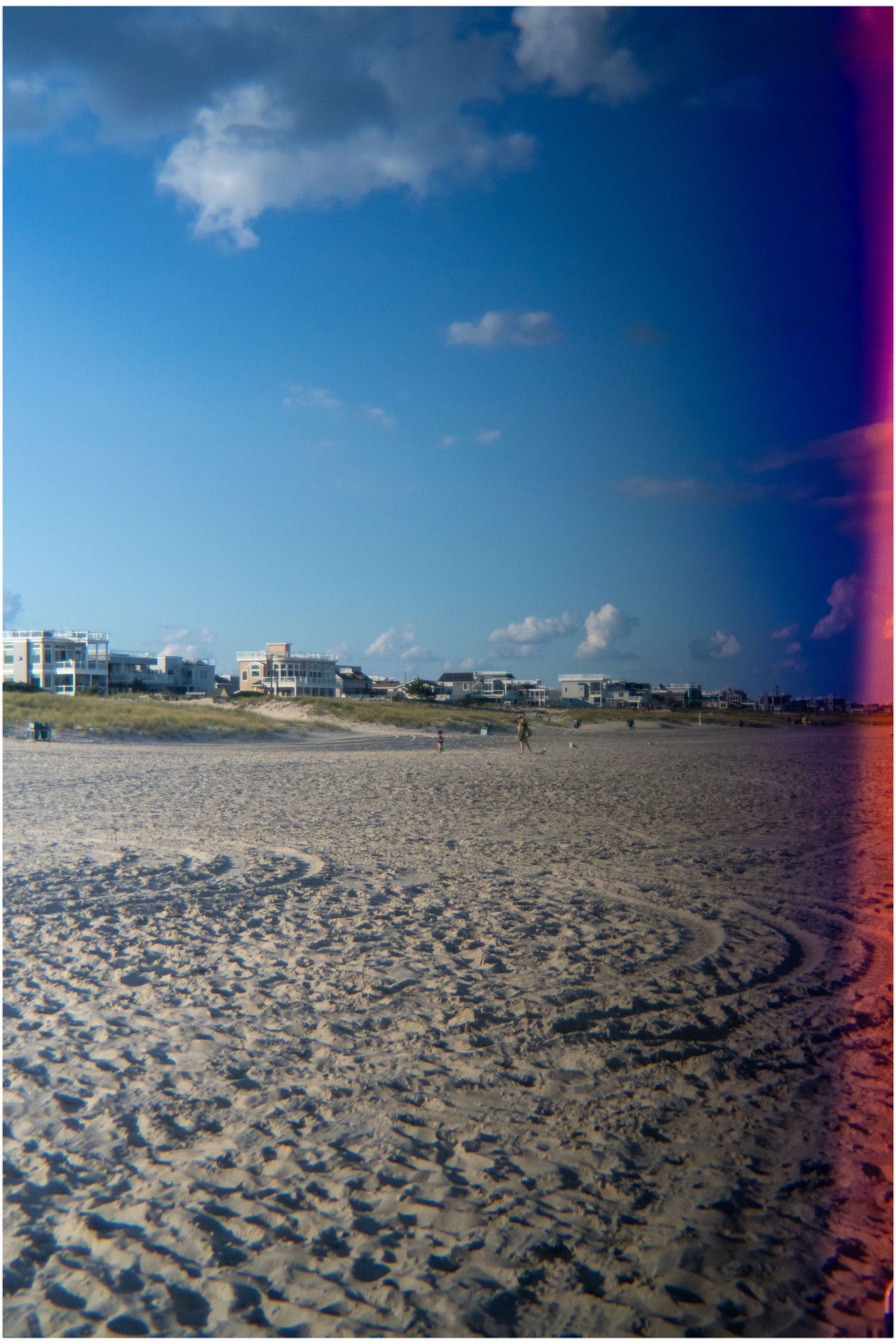 Sandy beach with footprints leading to beachfront houses under a blue sky with scattered clouds.