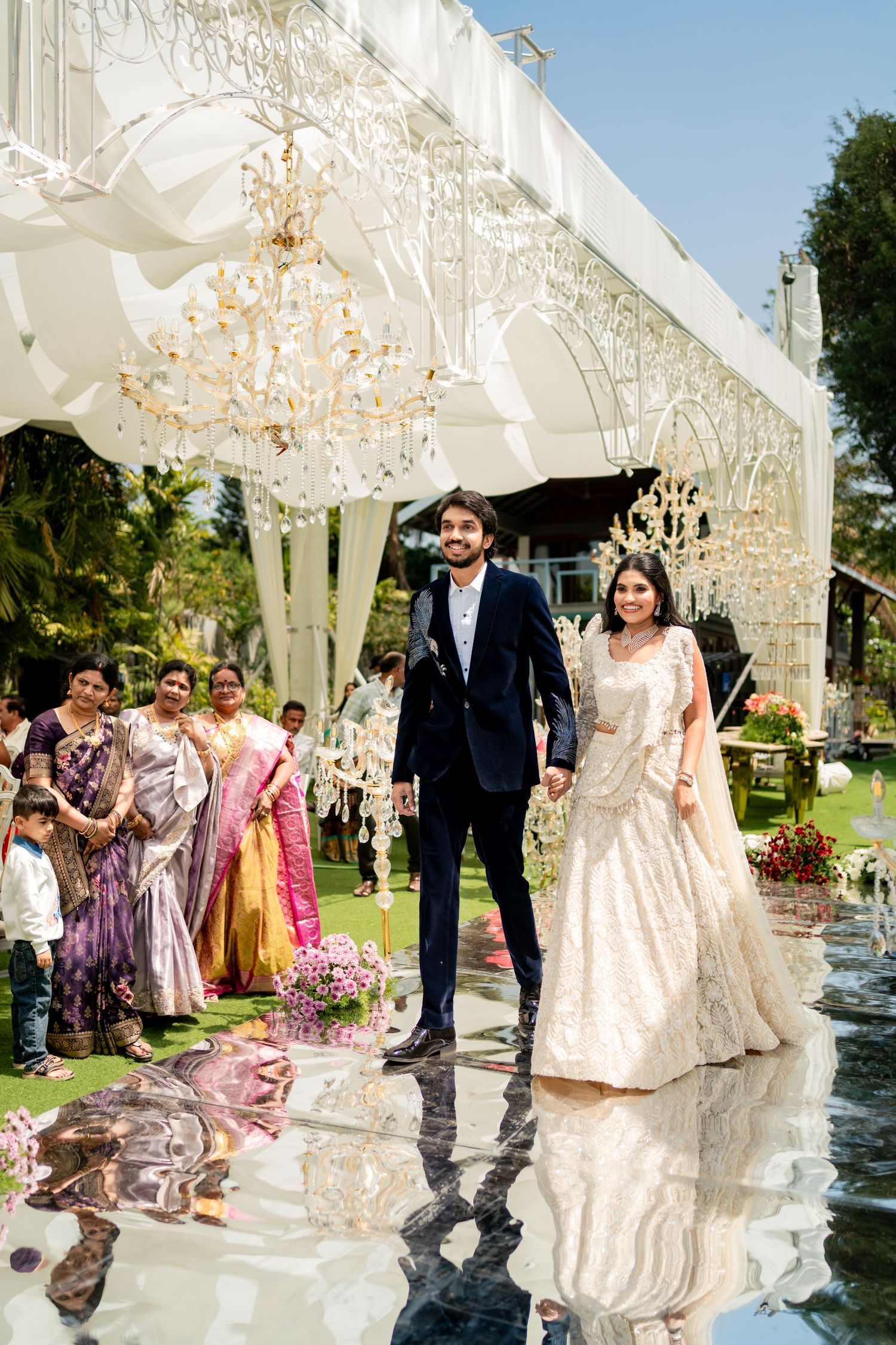 Indian bride and groom walking hand in hand on reflective floor under elegant chandeliers at outdoor wedding.