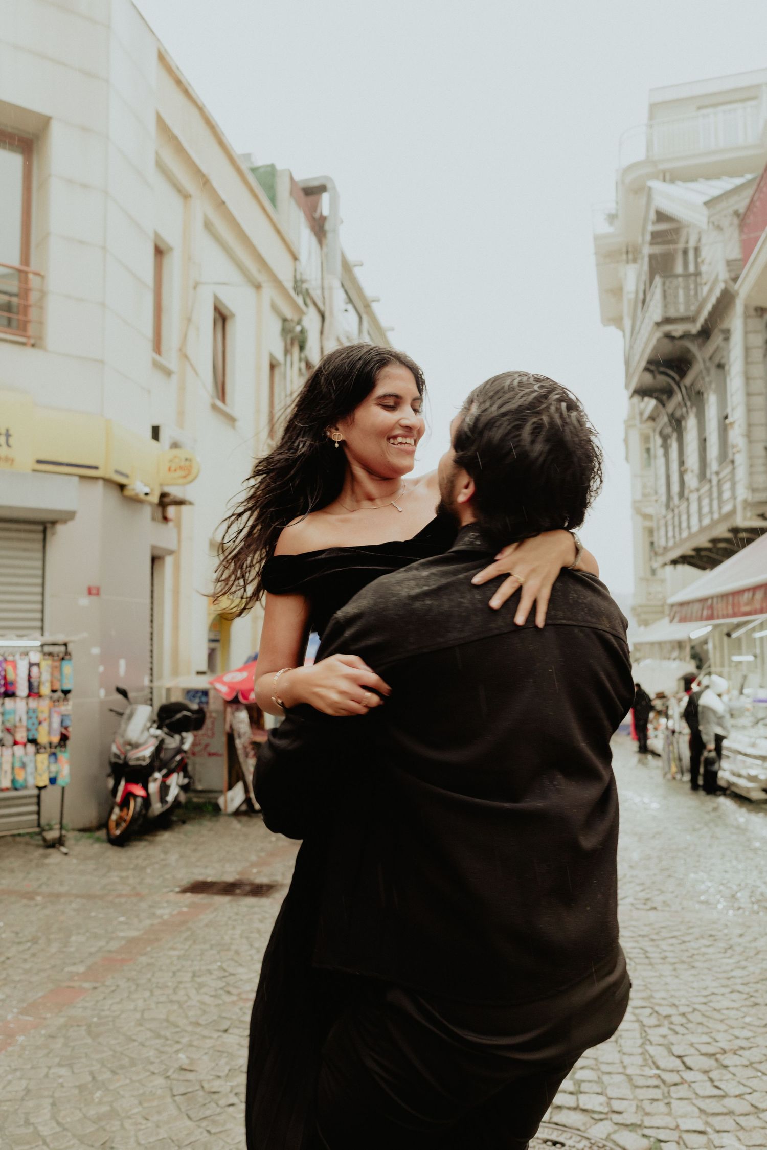 Couple in black outfits embrace and smile on a cobblestone street in Istanbul.