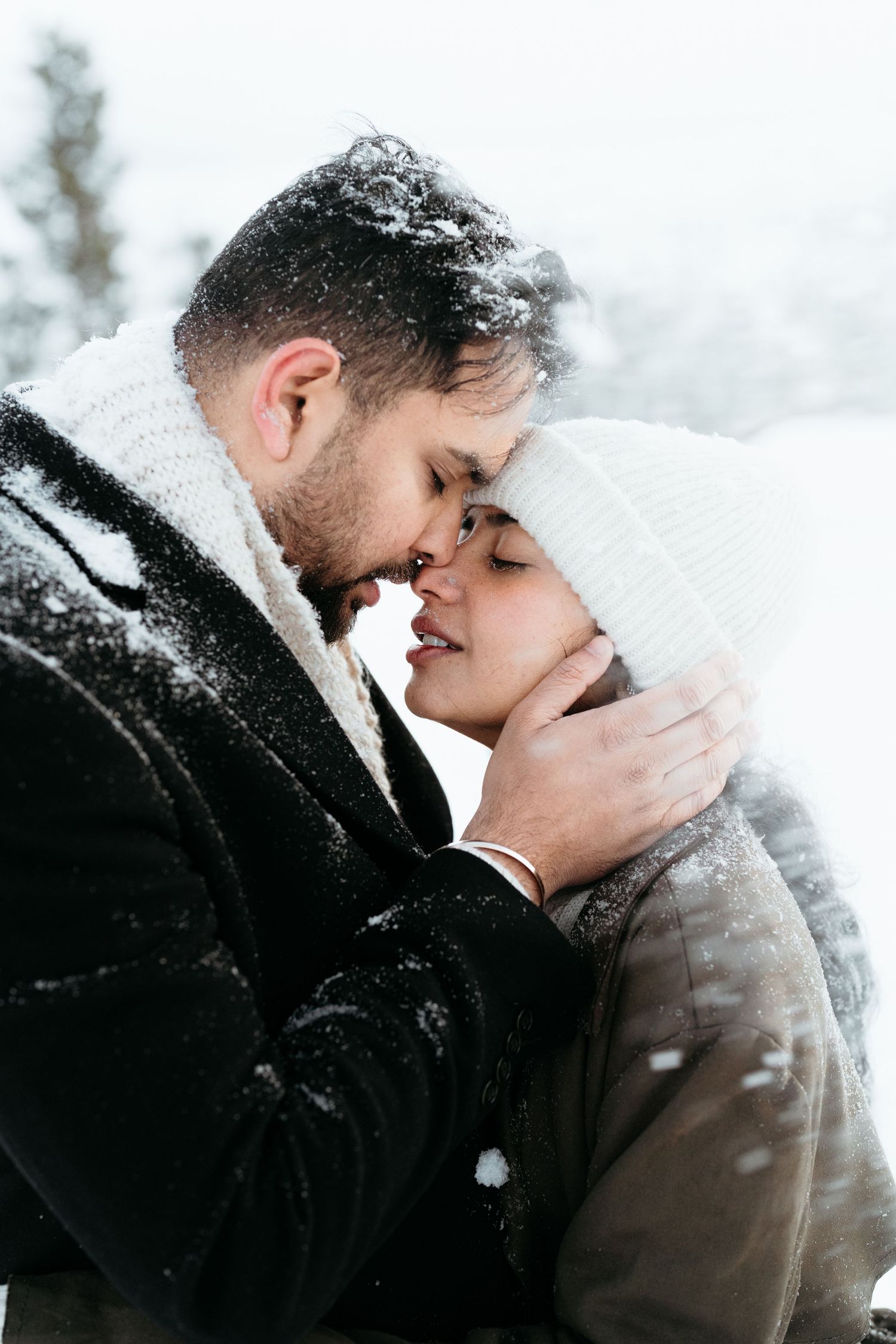 Romantic couple embracing in snowy winter landscape, man tenderly holding womans face as they lean in for a kiss.