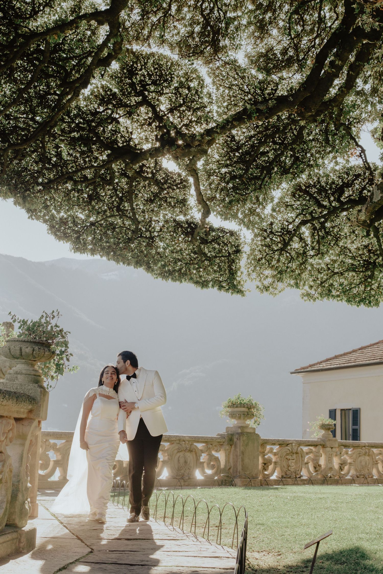 Bride and groom walk under a large tree at an elegant Italian villa with mountain views on their wedding day.
