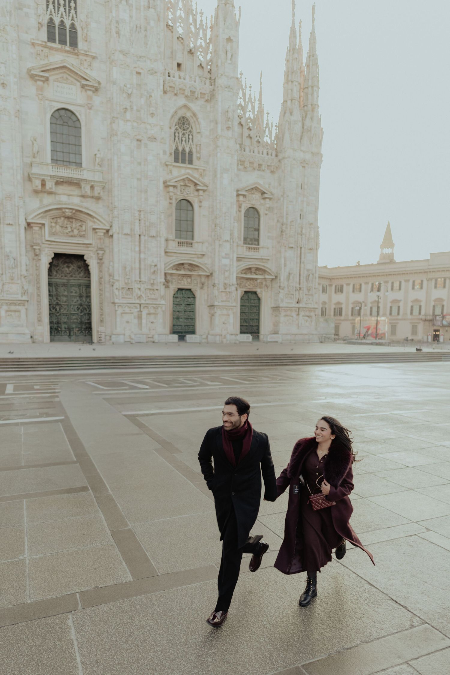 Couple running and laughing hand in hand across the plaza in front of Milan's Duomo.