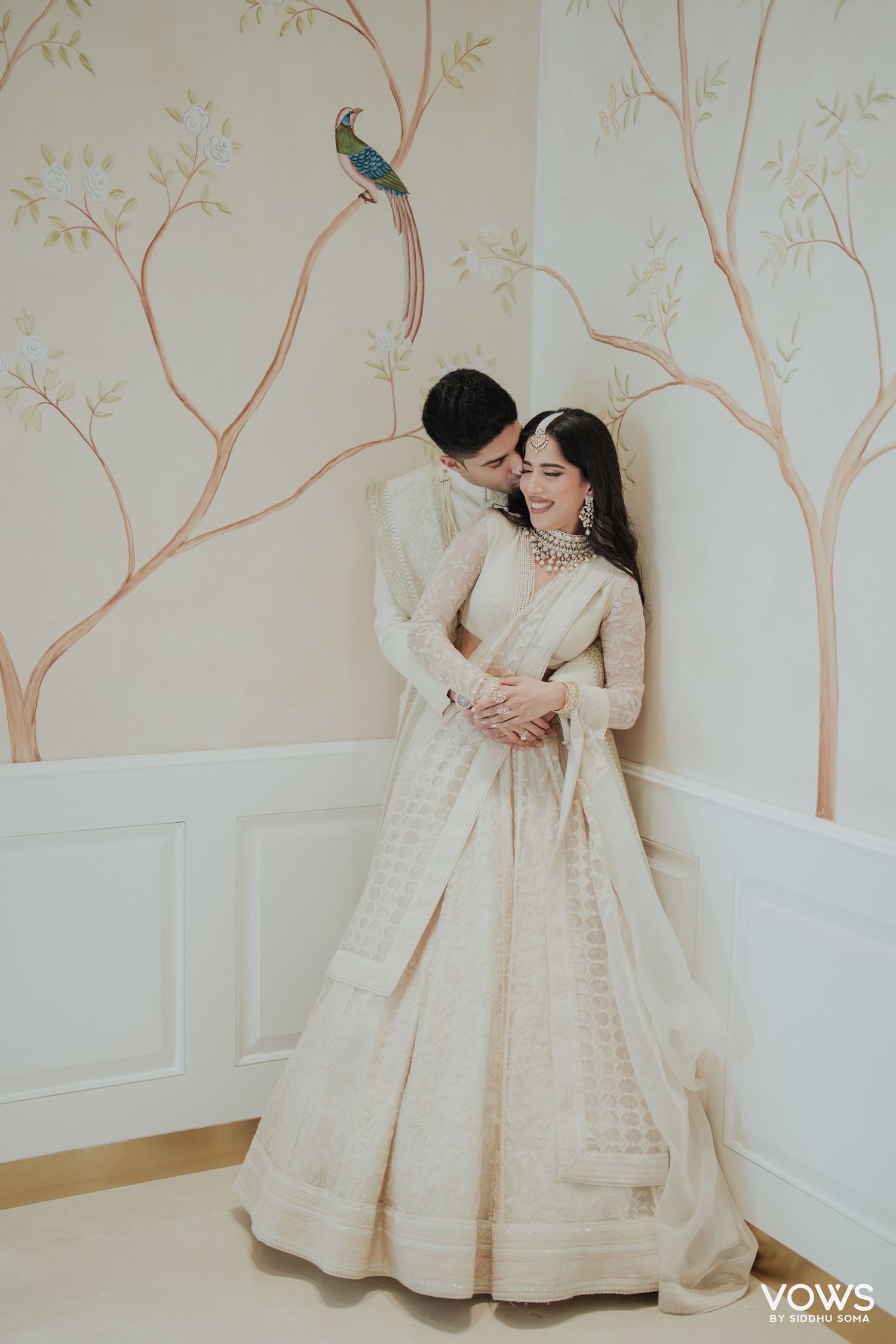 Indian bride and groom in ivory outfits pose romantically against a wall with floral mural backdrop.