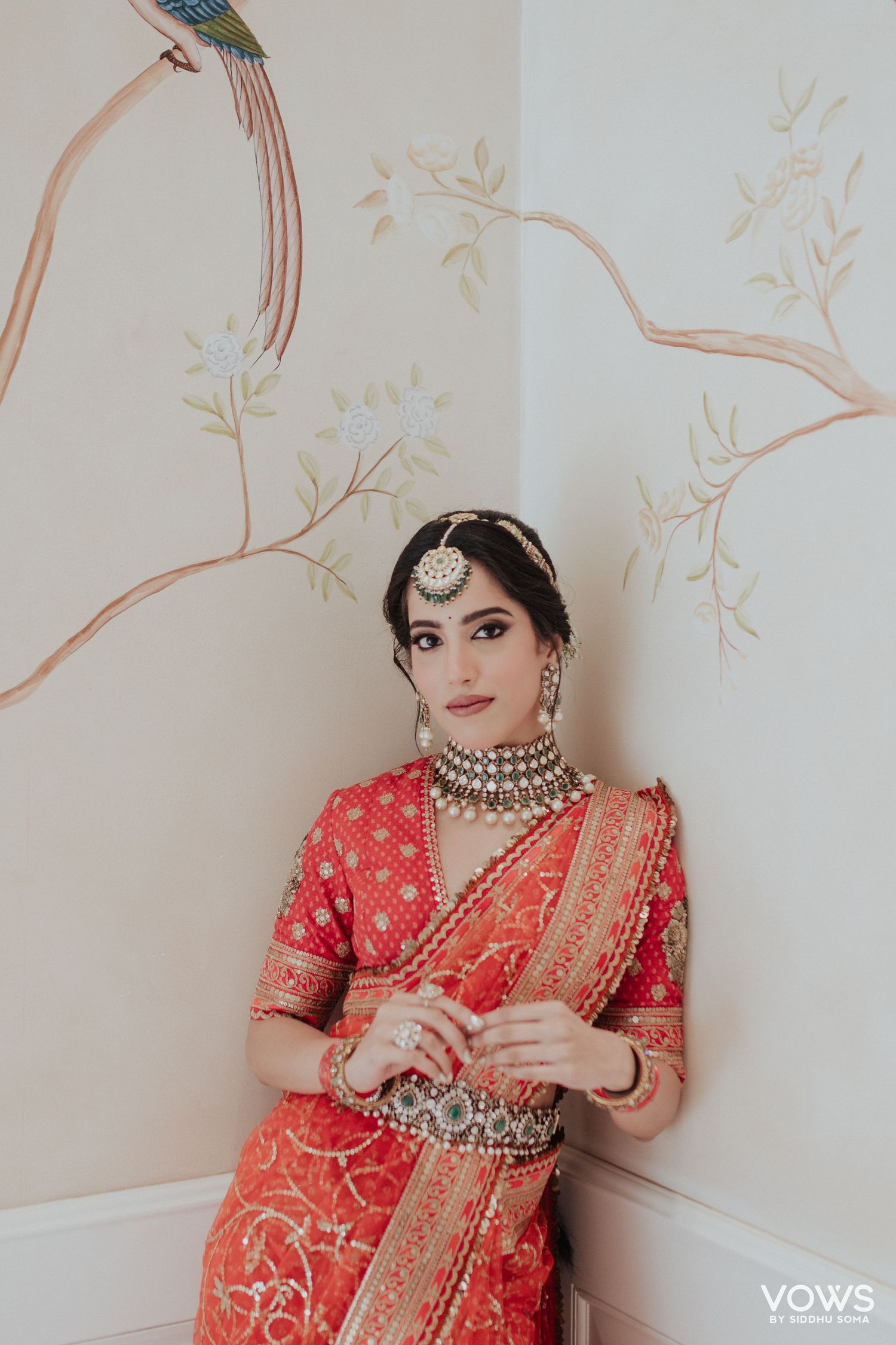 Bride in red embroidered saree with silver choker necklace and maang tikka poses against floral mural wall.