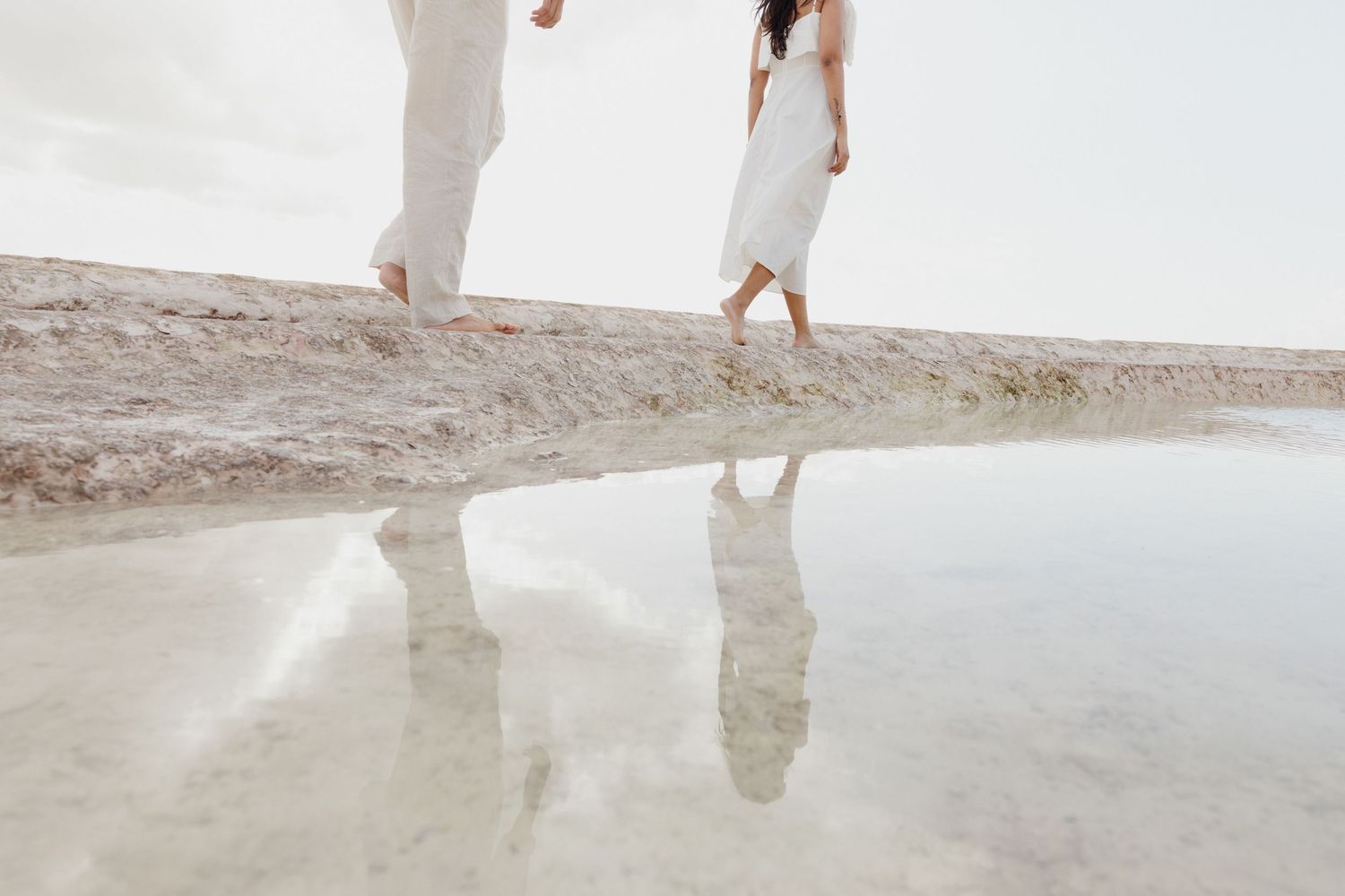 Couple in white attire walking barefoot on a sandy shore with a serene water reflection below.