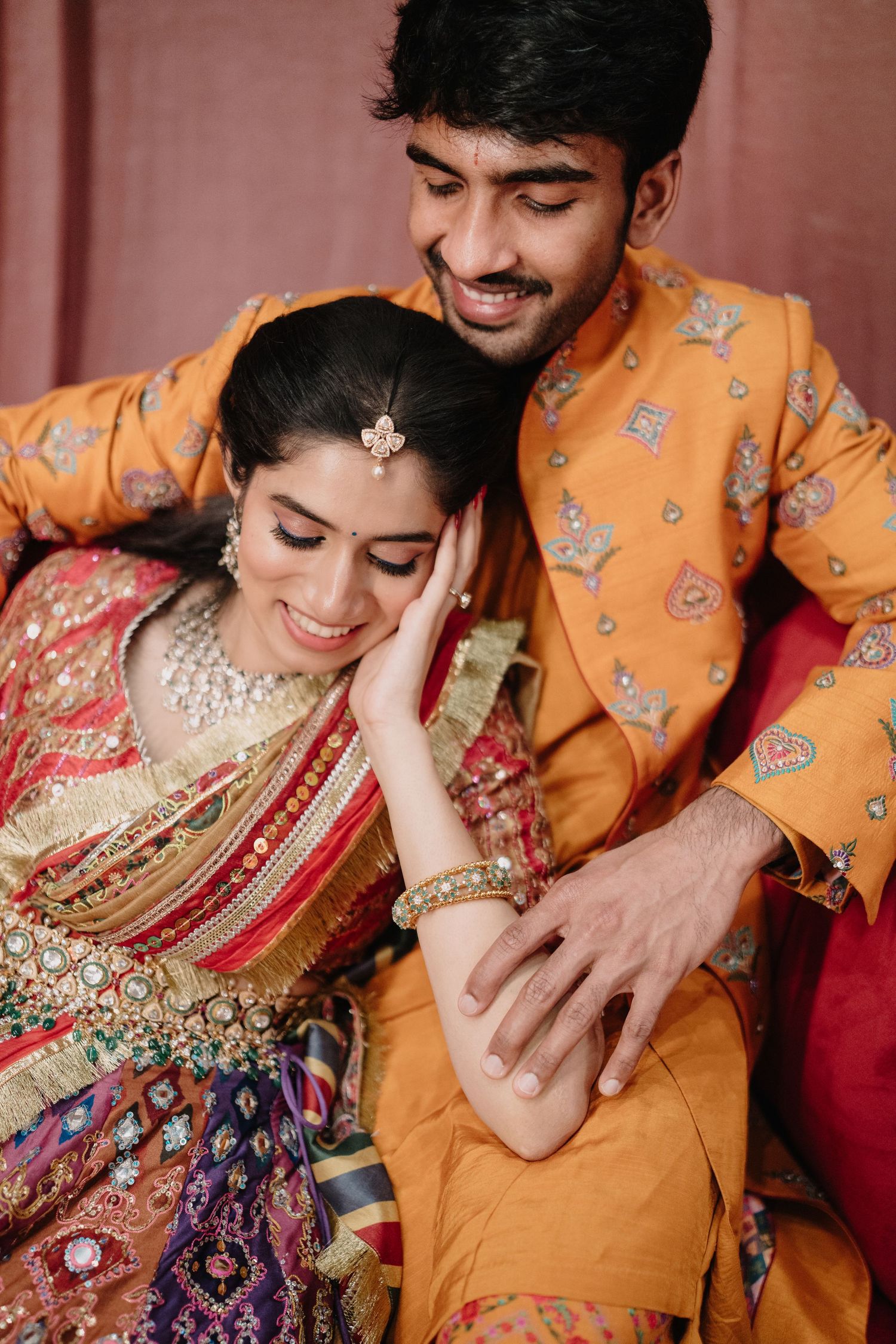A smiling Indian couple in traditional attire, bride in colorful lehenga and groom in orange sherwani, posing intimately.