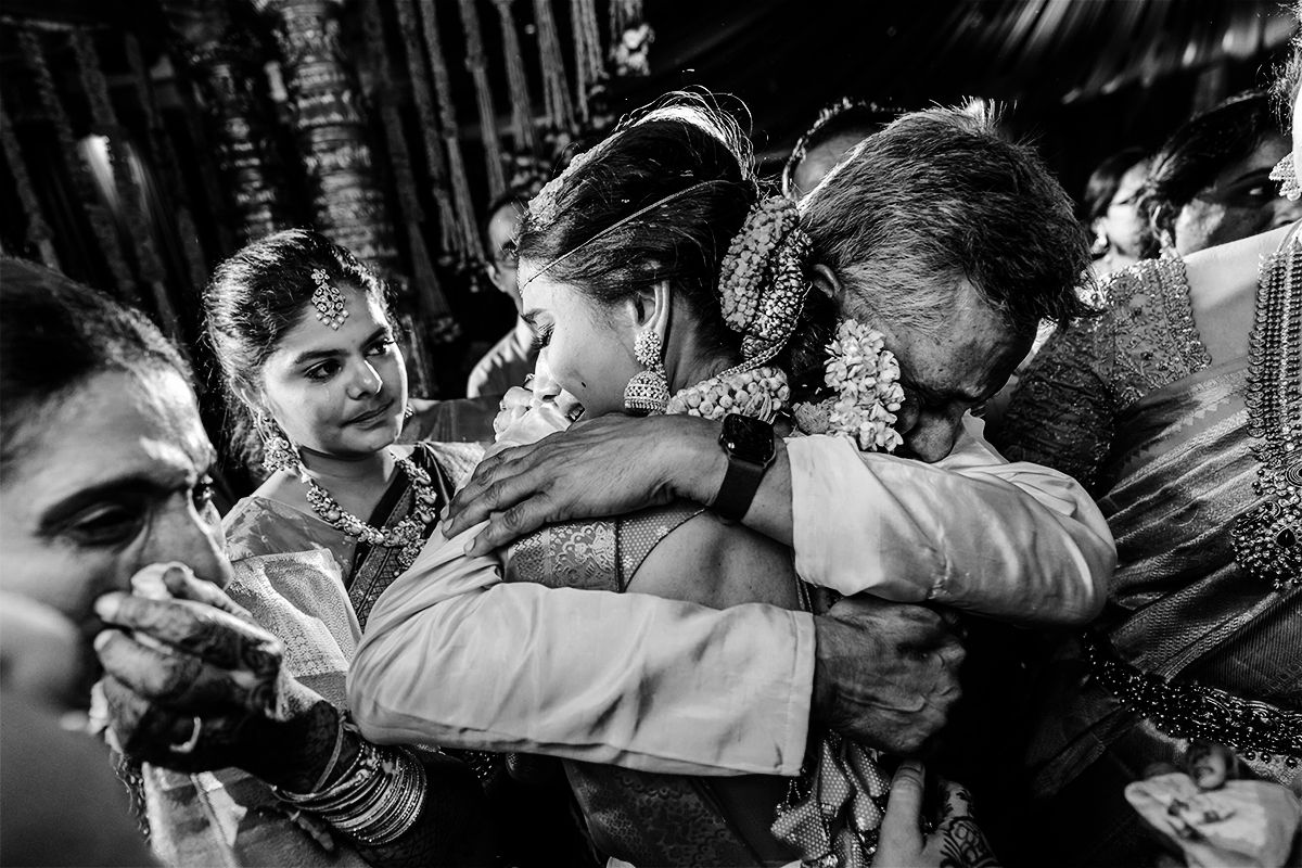 Black and white photo of emotional family embrace at traditional Indian wedding ceremony with floral decorations.
