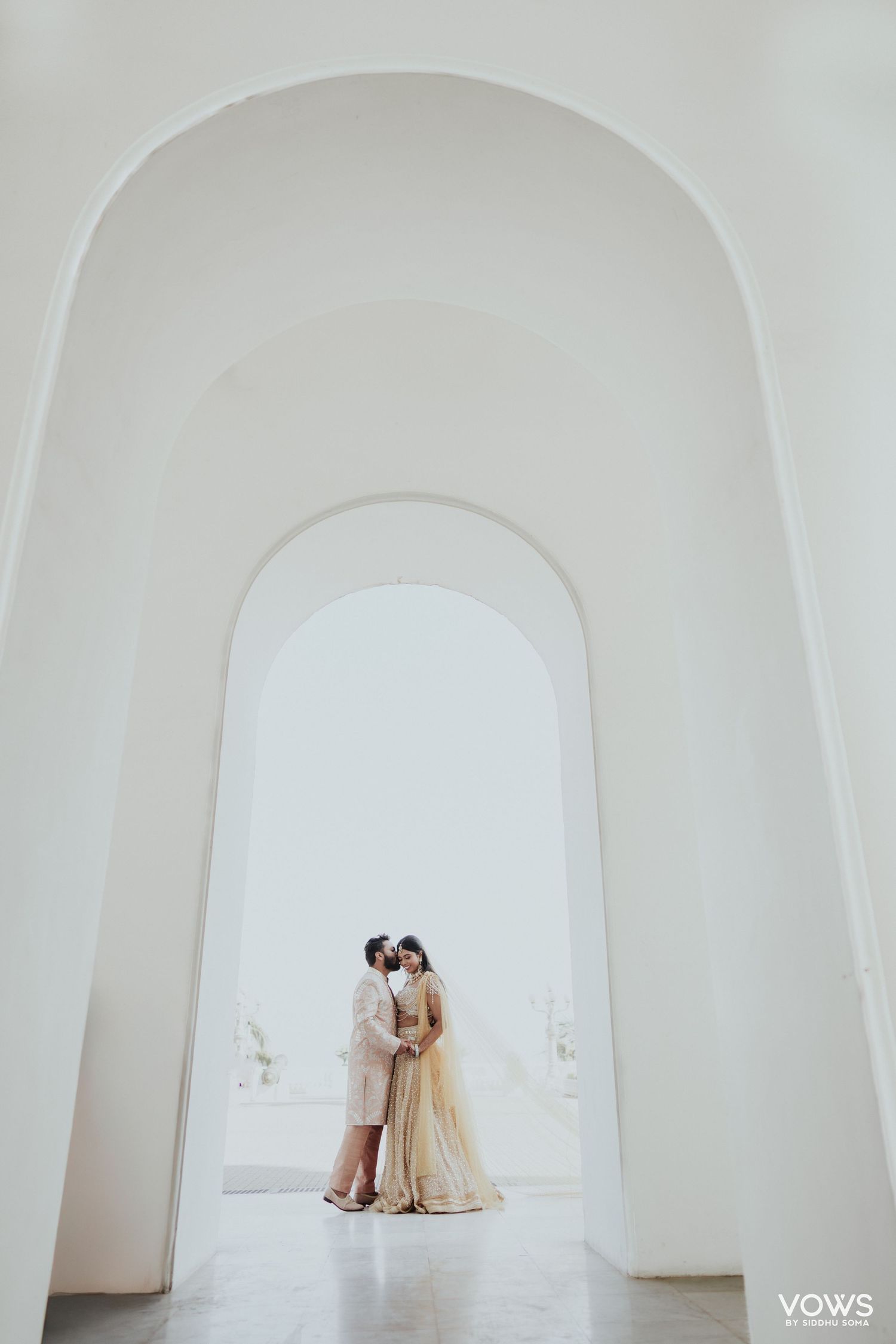Couple sharing a romantic moment framed by white arched hallway in traditional Indian wedding attire.