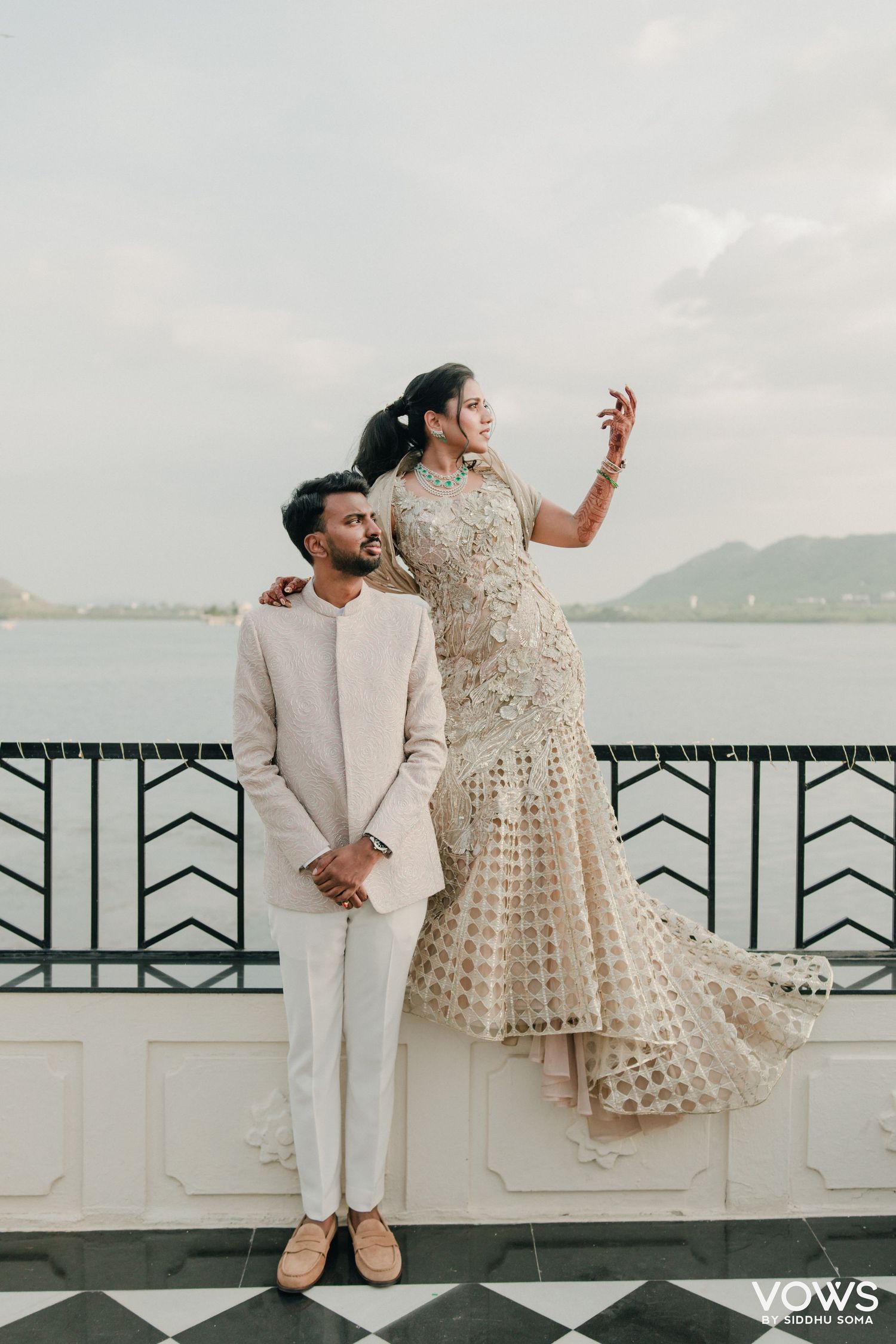 Indian couple in elegant wedding attire pose on a lakeside terrace, bride in gold gown stands on railing.