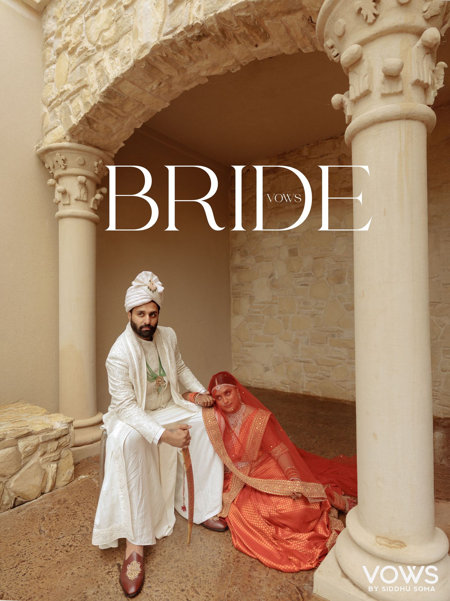 Indian couple in traditional wedding attire pose under stone archway, groom in white sherwani, bride in red saree.