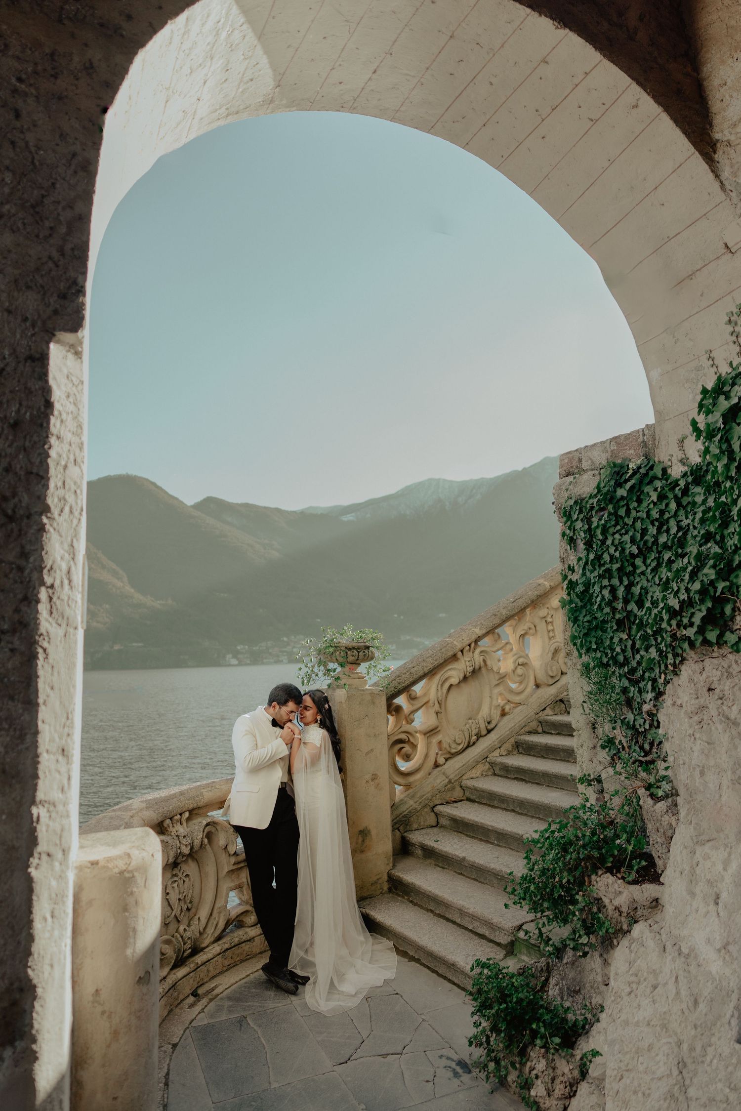 Bride and groom embrace under stone arch at Lake Como villa, with scenic mountain backdrop and ornate staircase.
