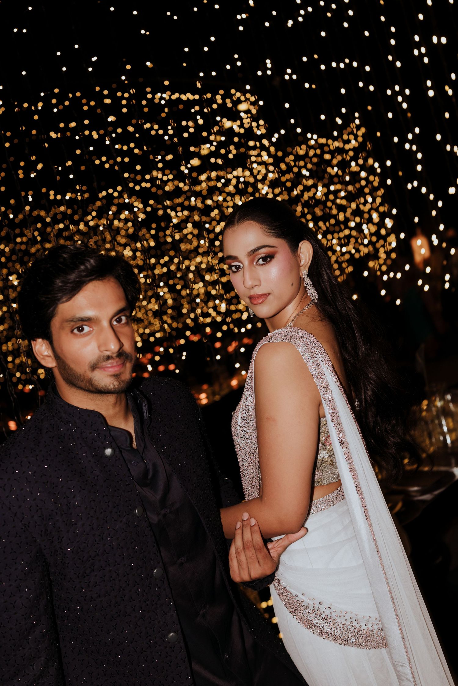 Couple in elegant Indian attire posing against a bokeh light backdrop at a festive night event.