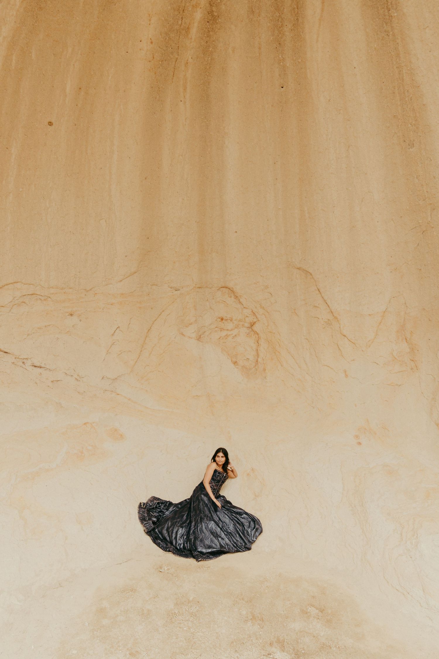Woman in a flowing black gown standing against a vast sandy desert wall, creating a dramatic contrast.