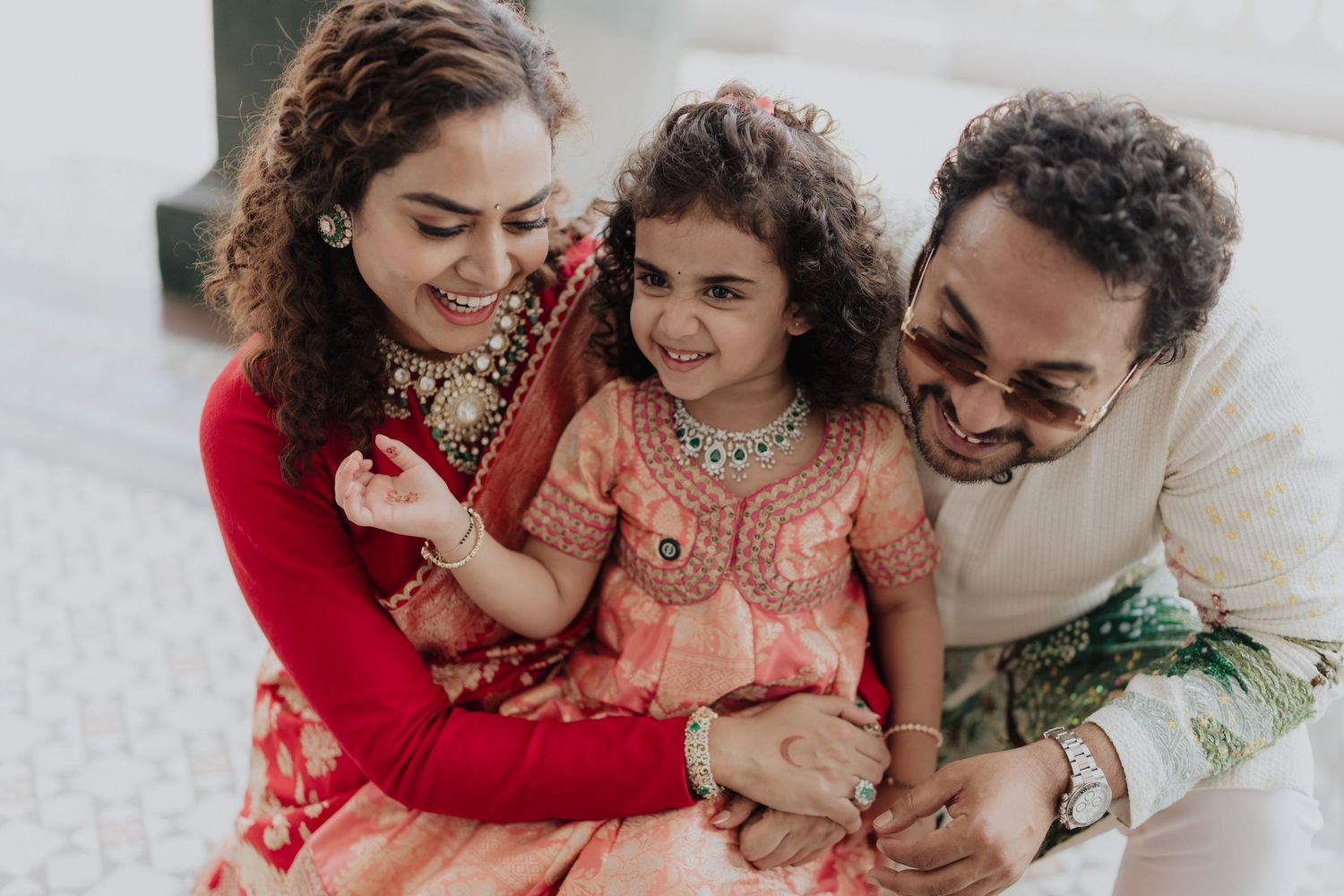 Smiling family in traditional Indian attire embracing, with a young girl in a peach embroidered outfit seated between her mother and father.