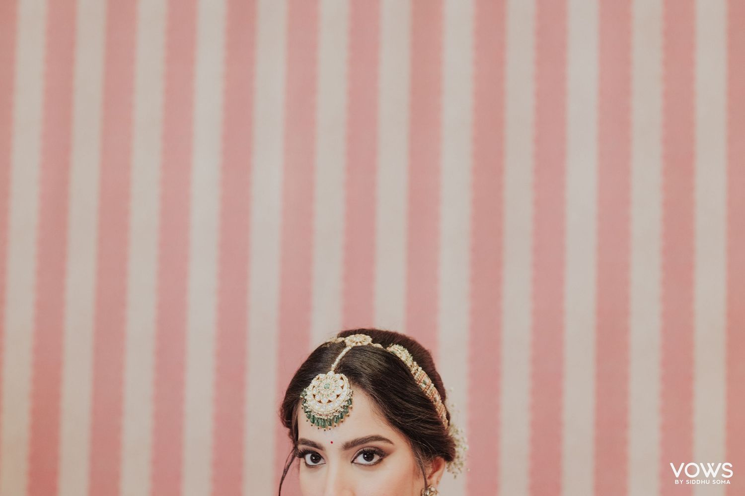 Indian bride in traditional jewelry with maang tikka posing against a pink and white striped background.