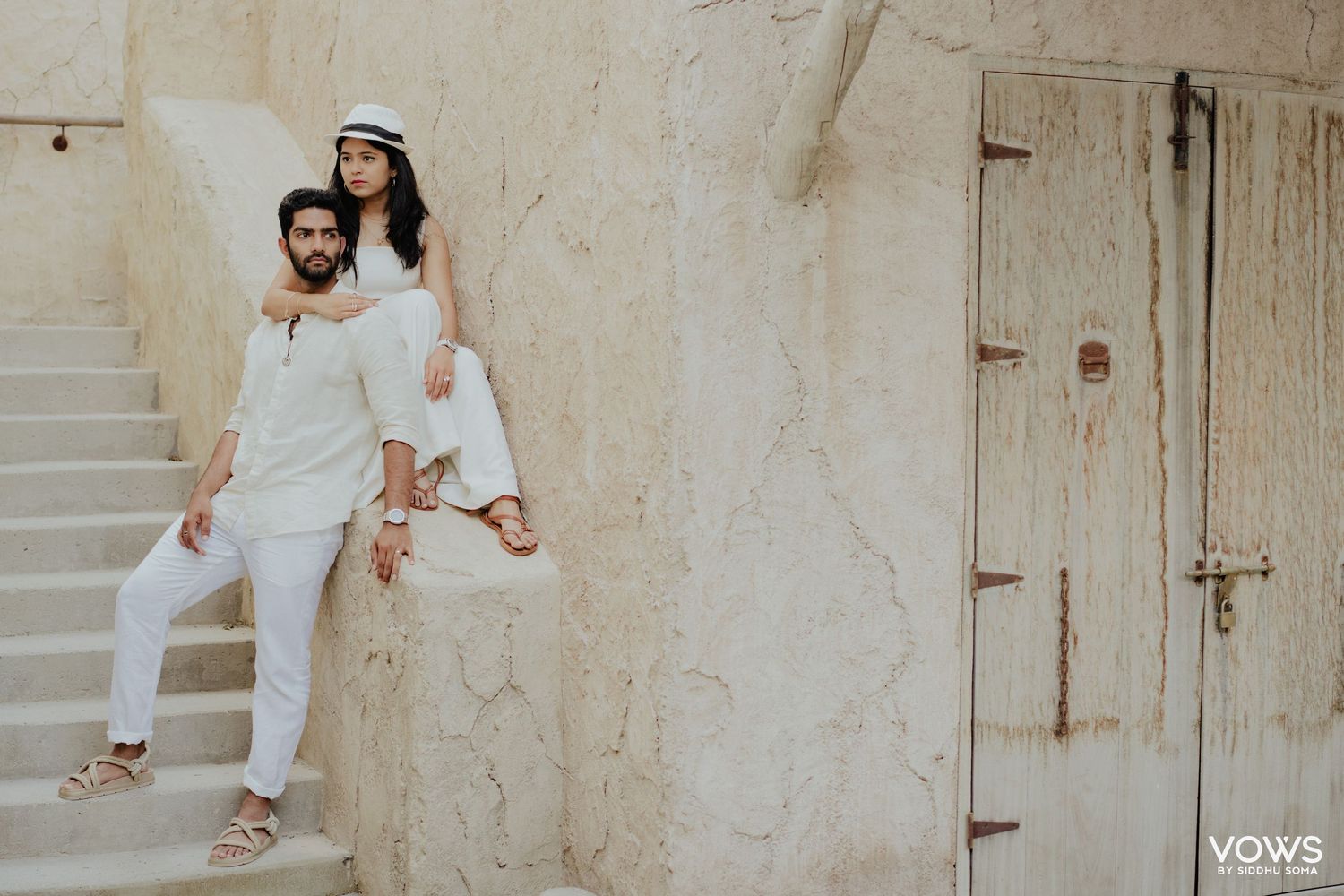 Couple in white outfits posing on rustic stone steps near a weathered door for a pre-wedding shoot.