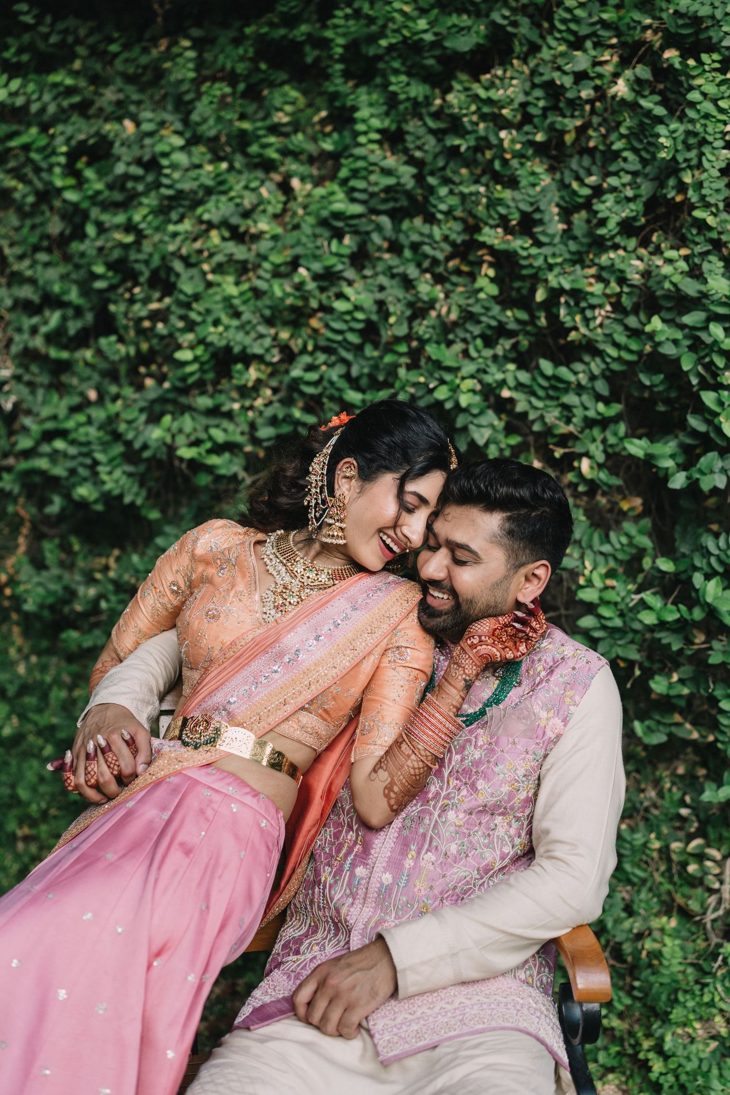Indian bride and groom in traditional attire share a joyful moment seated before a lush green ivy wall.