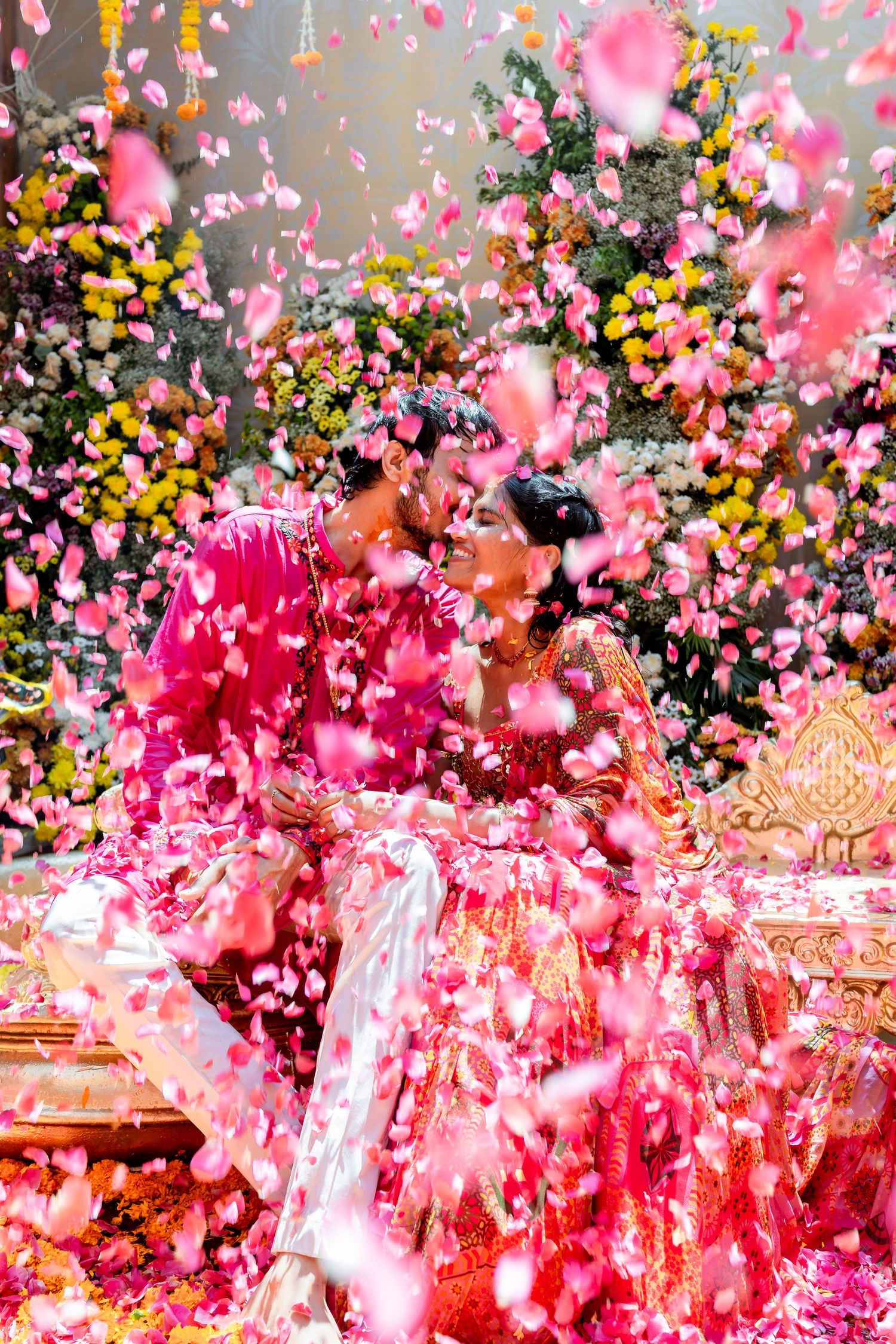 Bride and groom kissing surrounded by falling pink rose petals at vibrant Indian wedding.