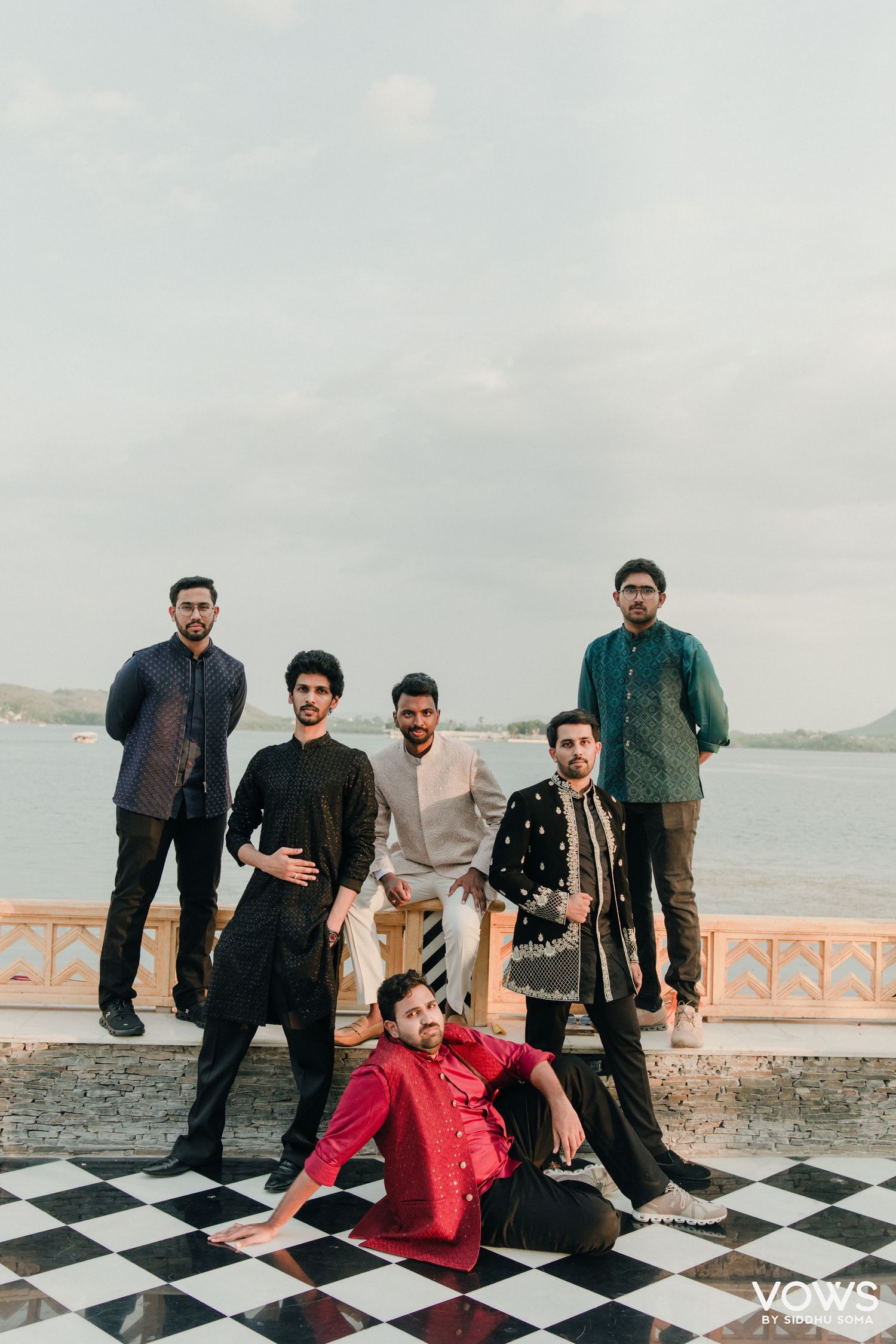 Six men in traditional Indian attire pose on a checkered floor by a lakeside at a wedding celebration.