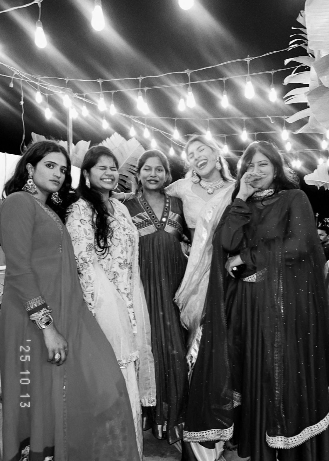 Five women in traditional Indian attire pose together under string lights at a festive outdoor event.
