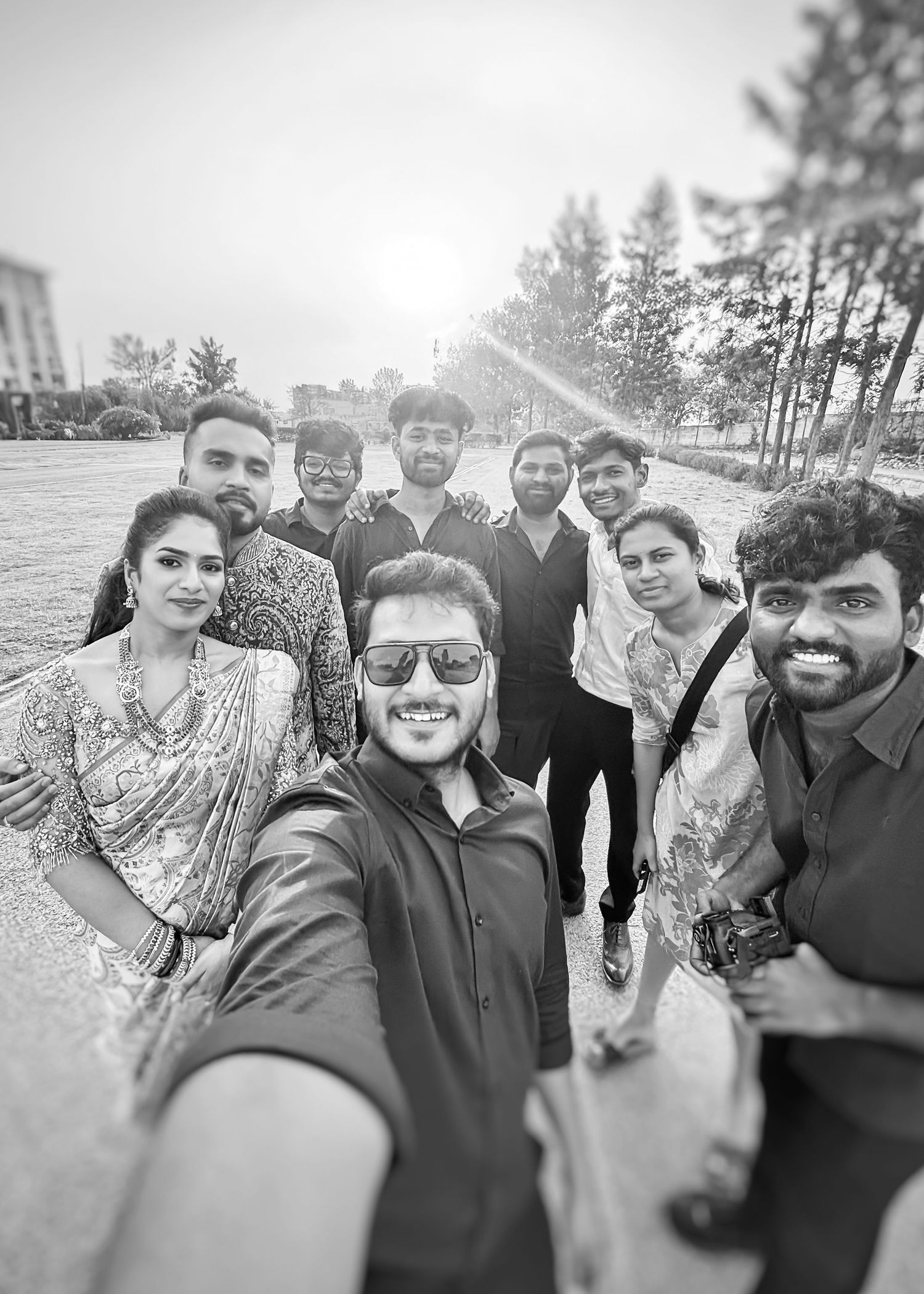 Black and white group selfie of friends posing outdoors, woman in saree among casually dressed men, trees in background.
